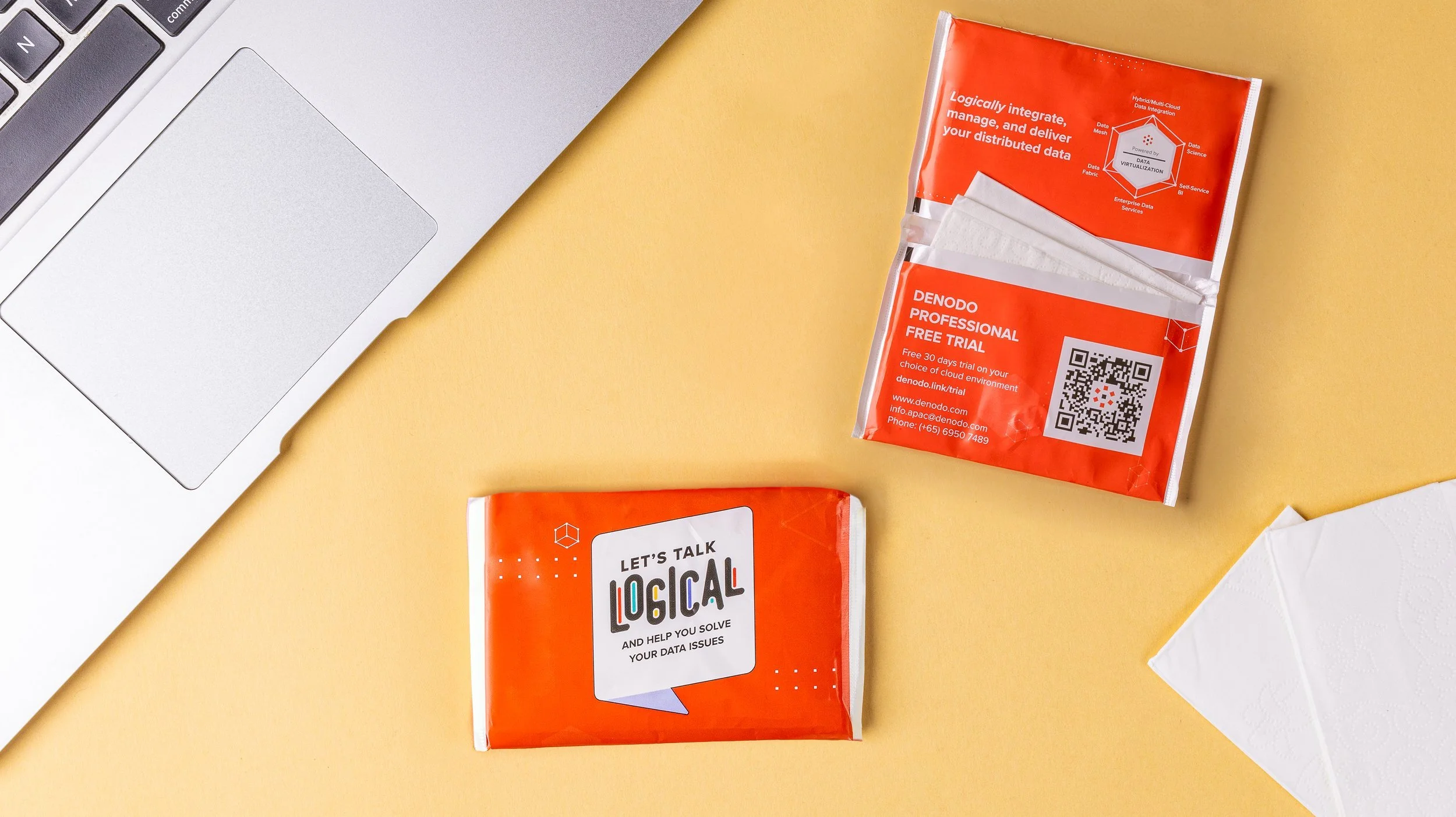 Denodo branded wet wipe sachets on a desk, one showing the Let's Talk Logical front, one the QR code reverse
