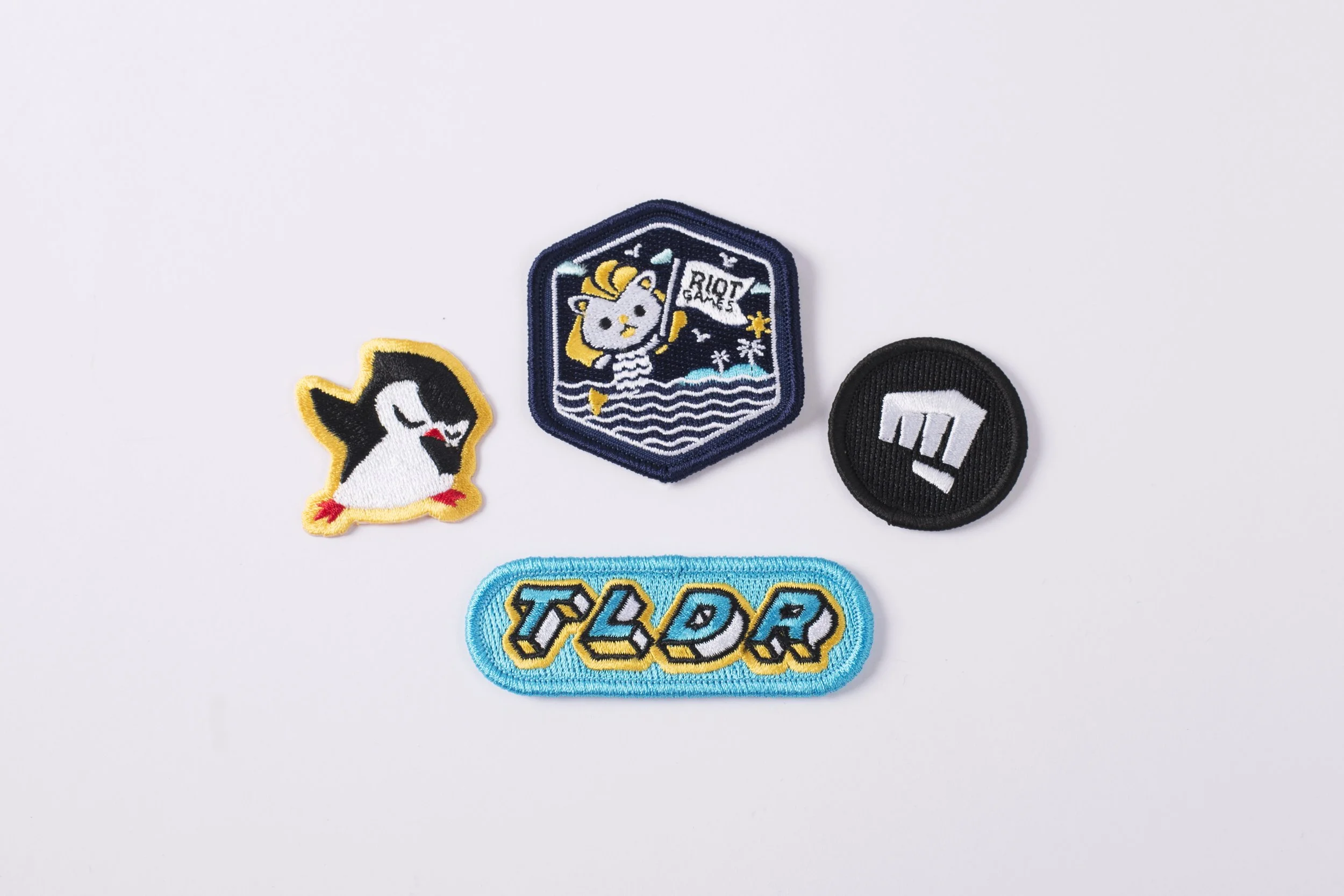 Four embroidered patches — a penguin, a hexagon crest, a fist logo and a TLDR badge — on white