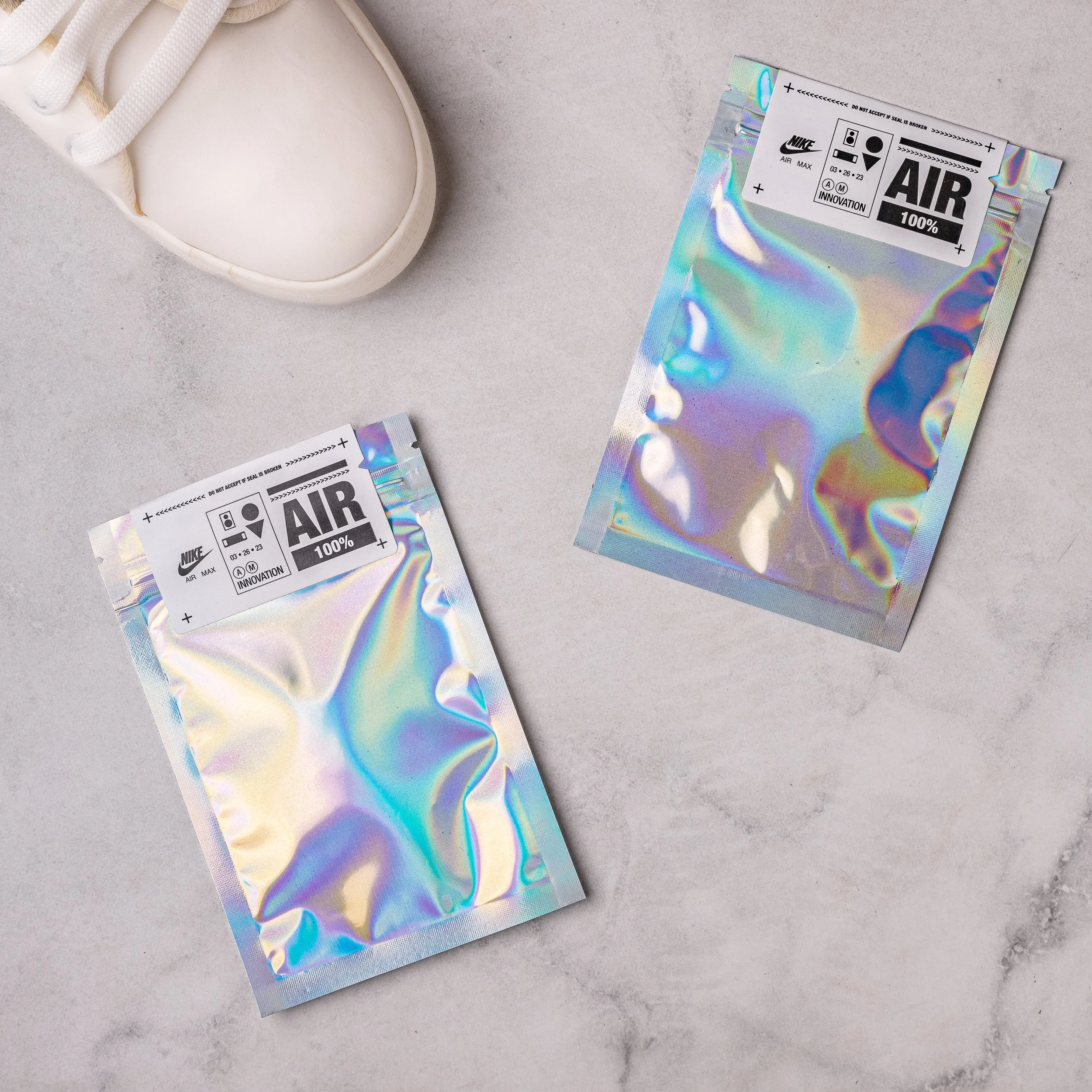 Two Nike Air Max holographic retail pouches laid beside a white sneaker on marble