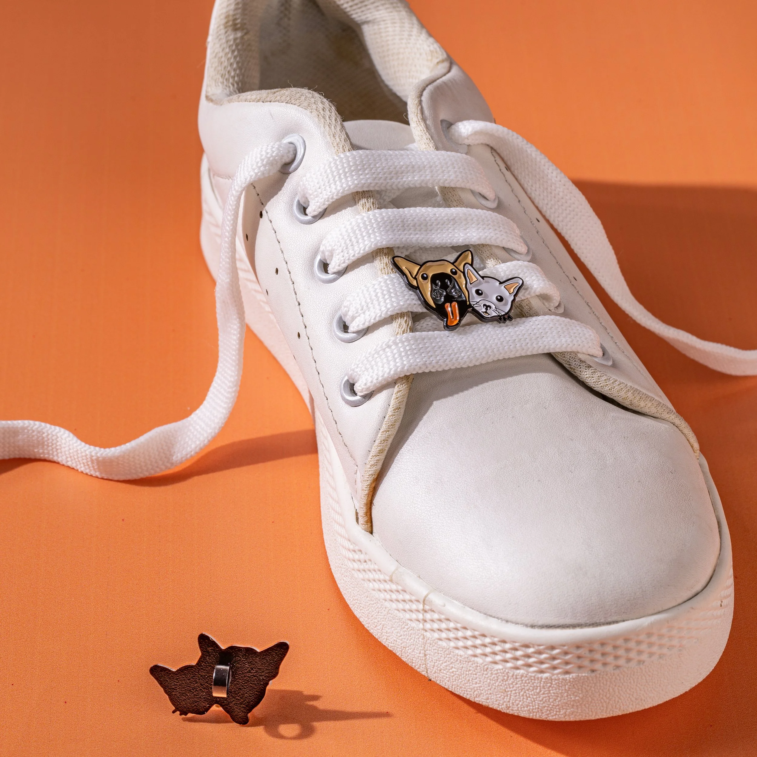 Enamel shoe charm of a dog and cat face fitted to a white sneaker, with the clip backing loose