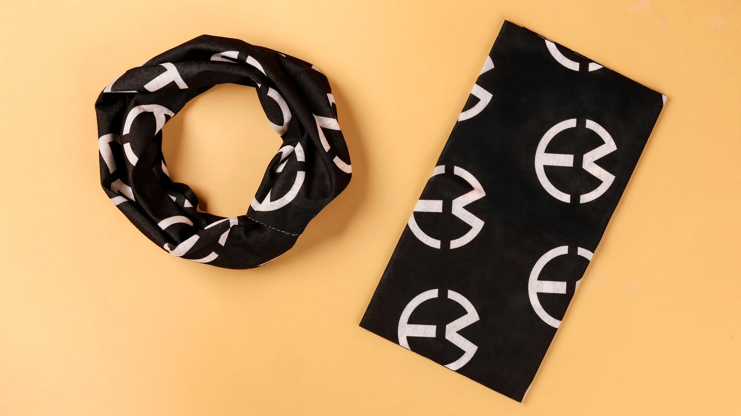 Black tube bandana printed with a repeating 
white circular logo, shown rolled and folded 
flat