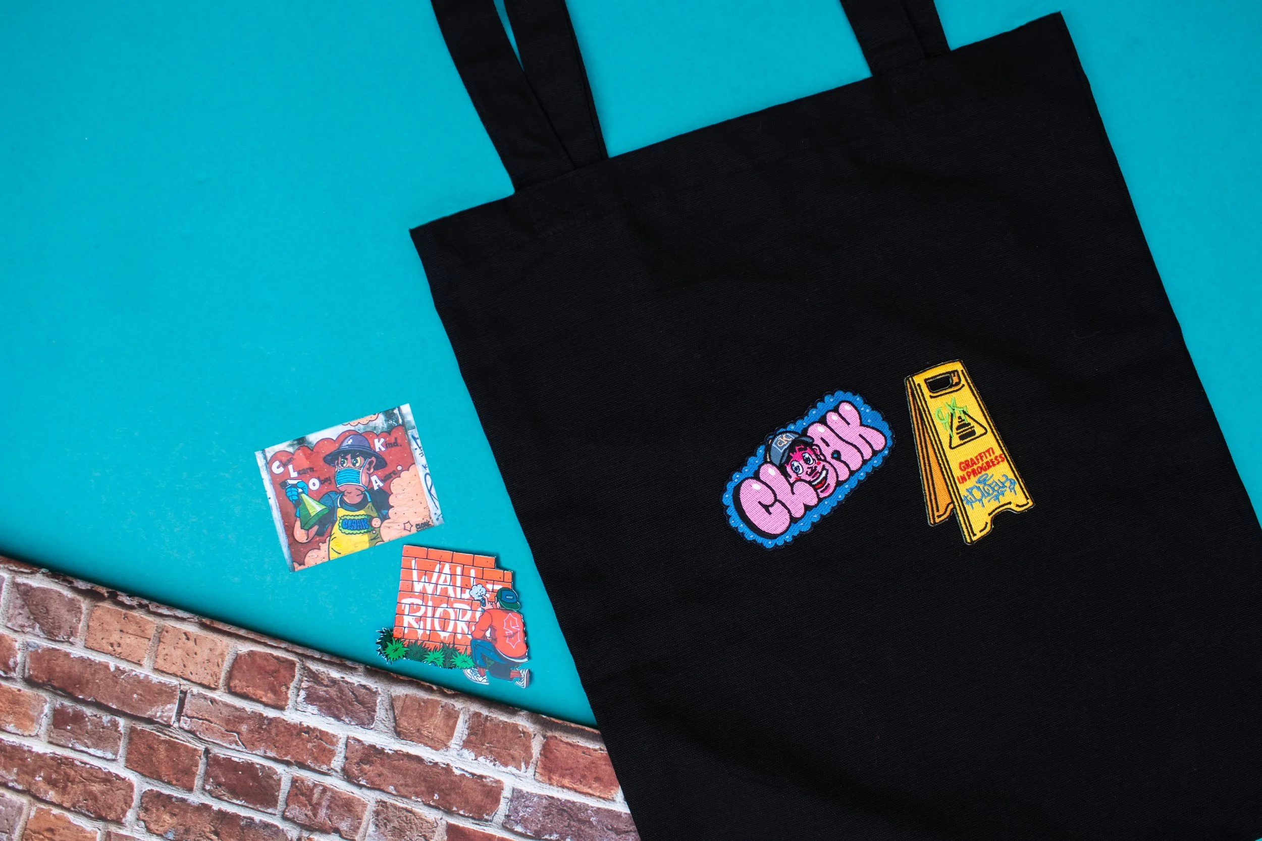 Cloak graffiti-style embroidered patches on a black tote bag with matching printed stickers