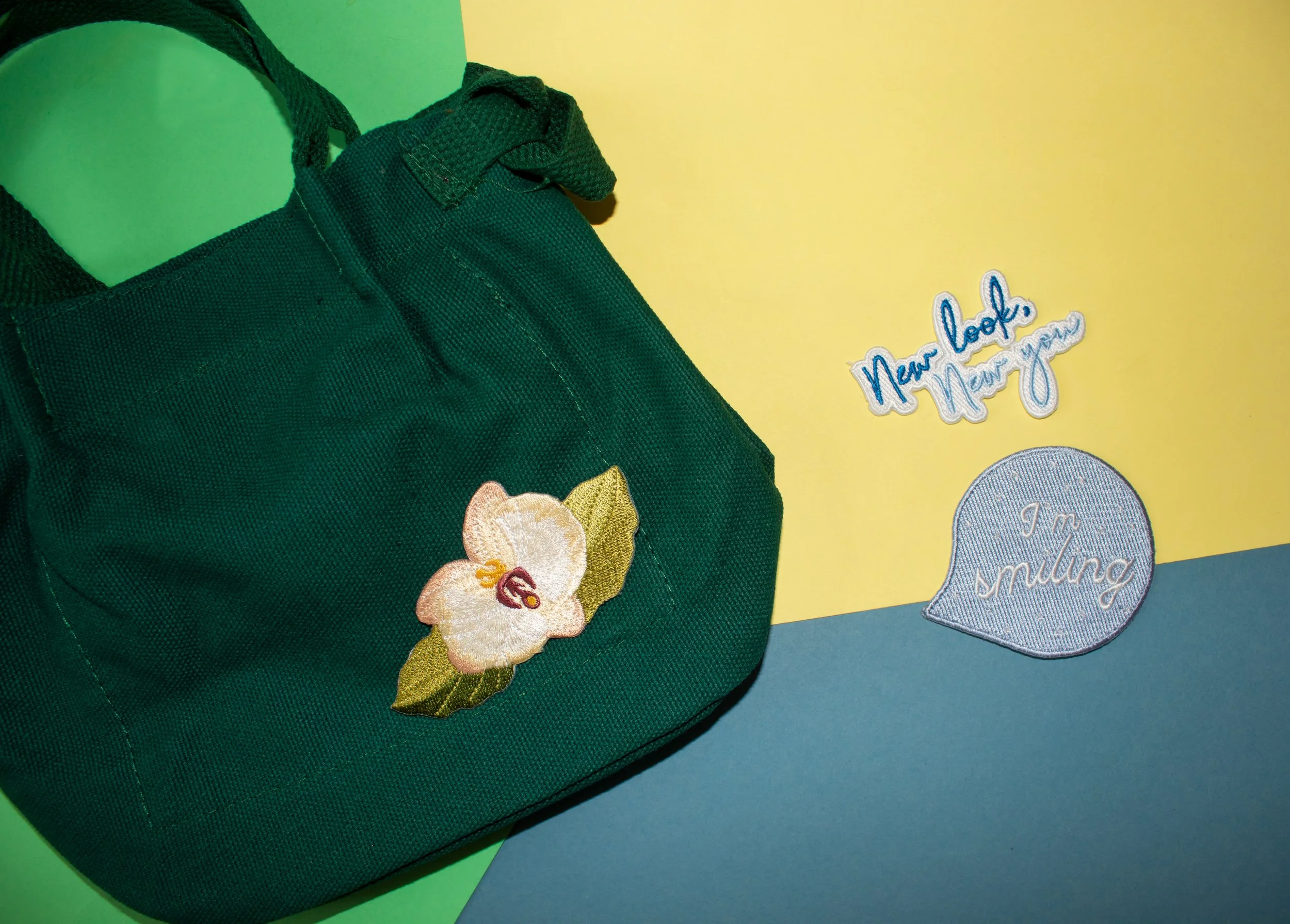 Embroidered orchid patch on a green tote bag beside New look New you and I am smiling script patches