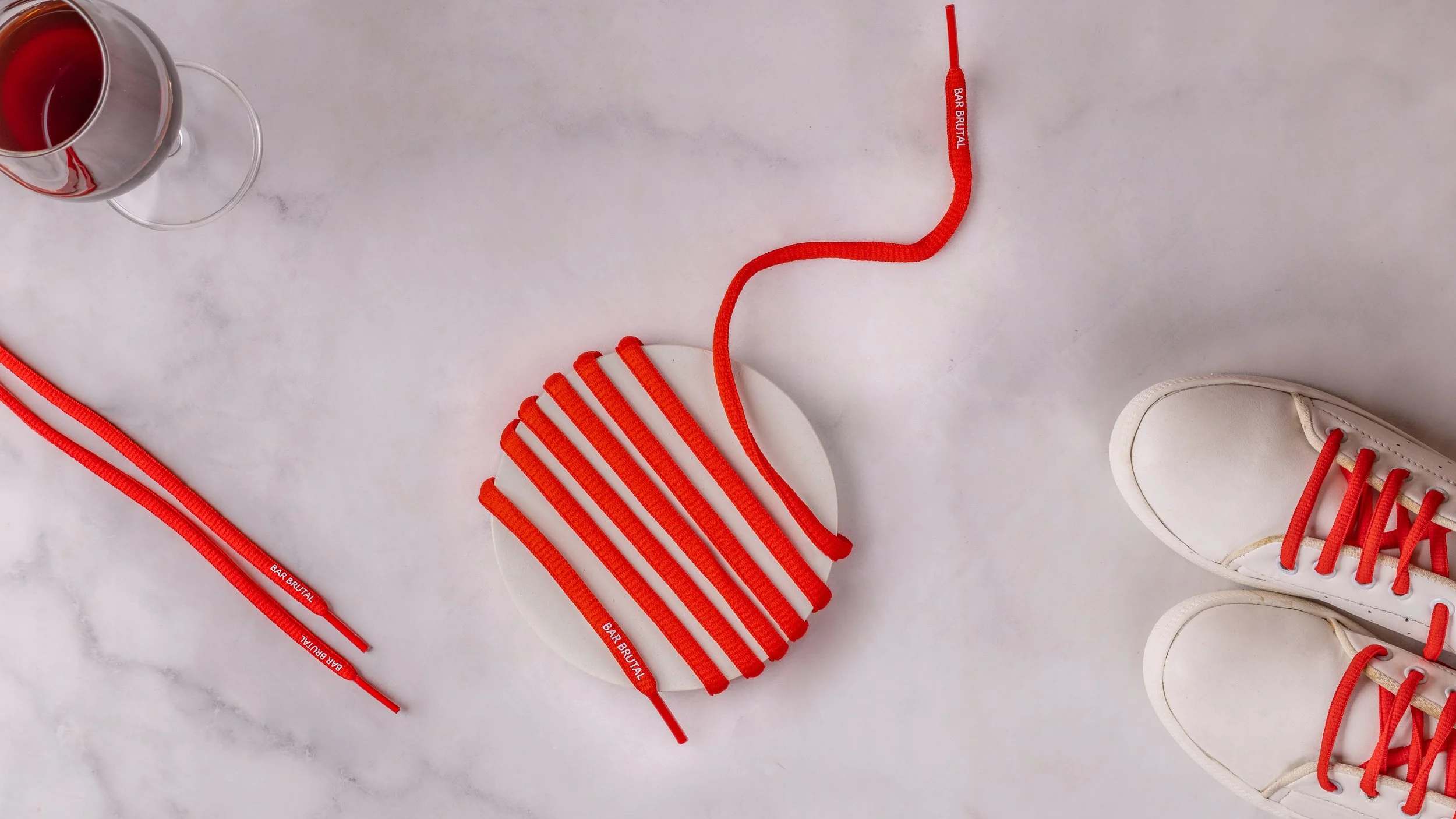 Red round shoelaces with a printed brand name of Bar Brutal, laced into white sneakers on a marble surface