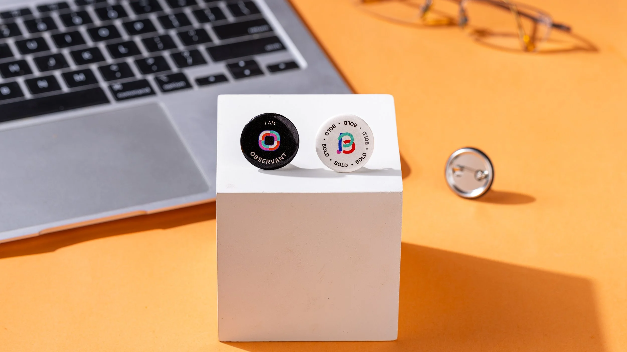 Black I Am Observant and 
white Bold slogan button 
badges on a white display 
cube beside a laptop and 
glasses