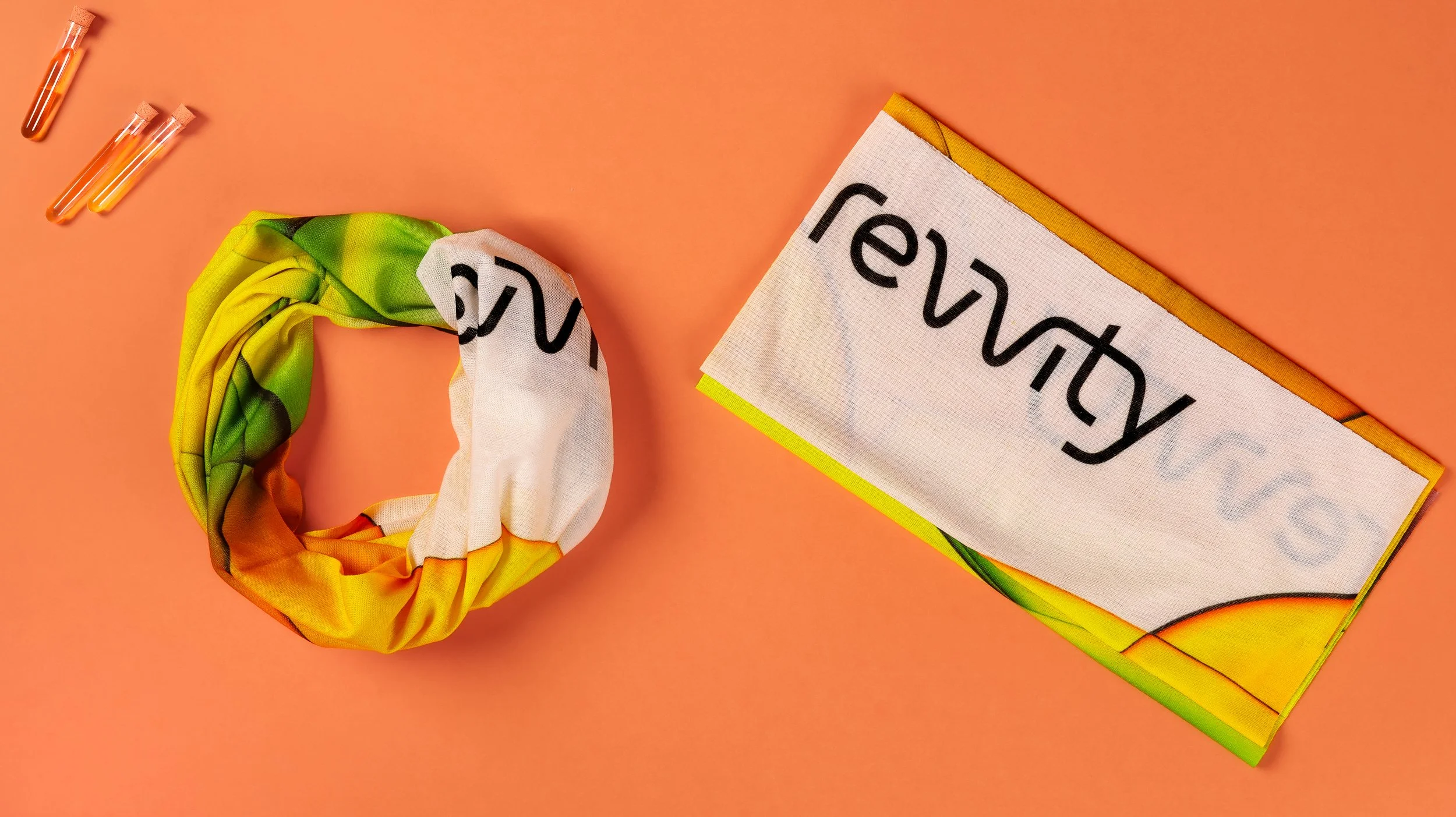 Revvity tube bandana in a green to orange 
gradient, shown rolled and folded flat beside 
test tubes