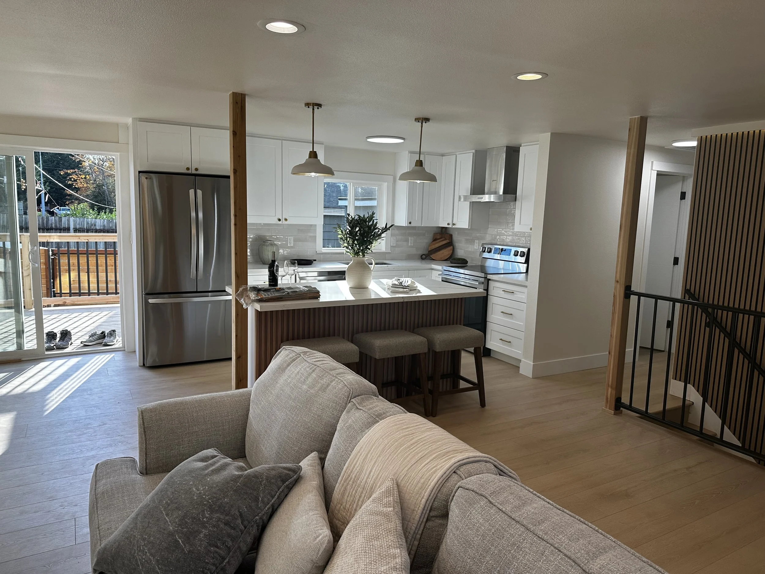 Open-concept kitchen and living area with beige sectional sofa, wooden flooring, stainless steel refrigerator, white cabinets, and pendant lighting over a kitchen island with a vase of flowers.