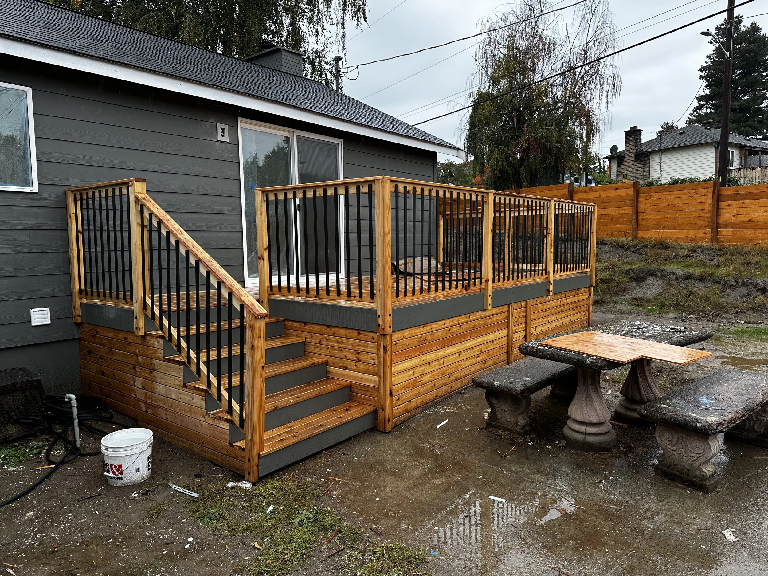 A house with gray siding and a black roof, featuring a newly built wooden deck with black metal railings. There is a sliding glass door leading to the deck and a window on the side of the house. In the background, other houses and trees are visible under an overcast sky. The ground in the yard is muddy with some puddles and scattered debris.