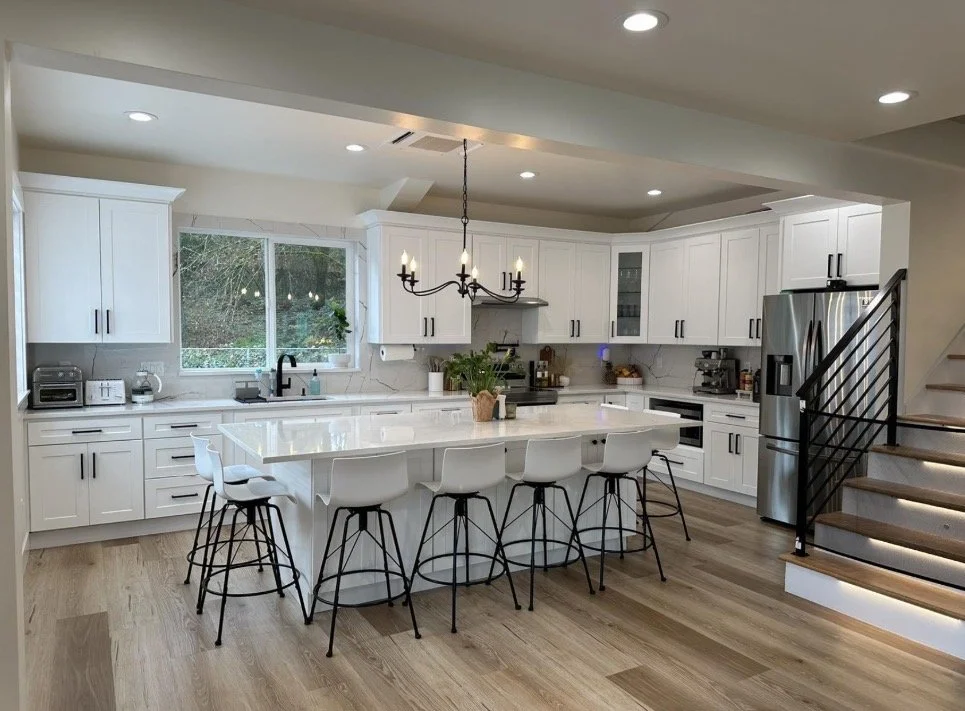 Modern white kitchen with an island, six white bar stools, stainless steel refrigerator, black railing staircase, and wooden floors