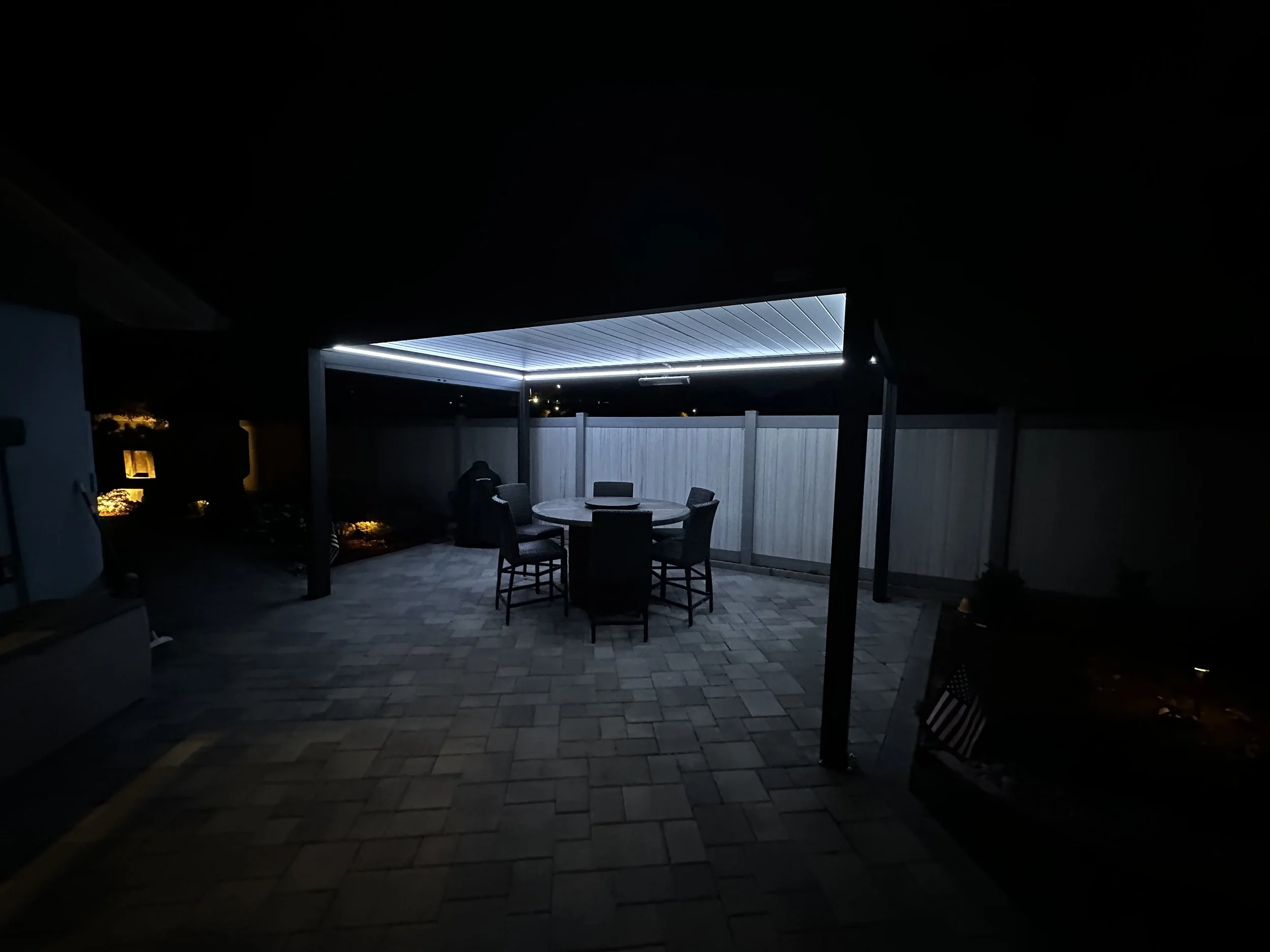 Nighttime outdoor patio with a round table and four chairs, enclosed by a light-colored fence, illuminated by blue LED lighting from a pergola overhead.