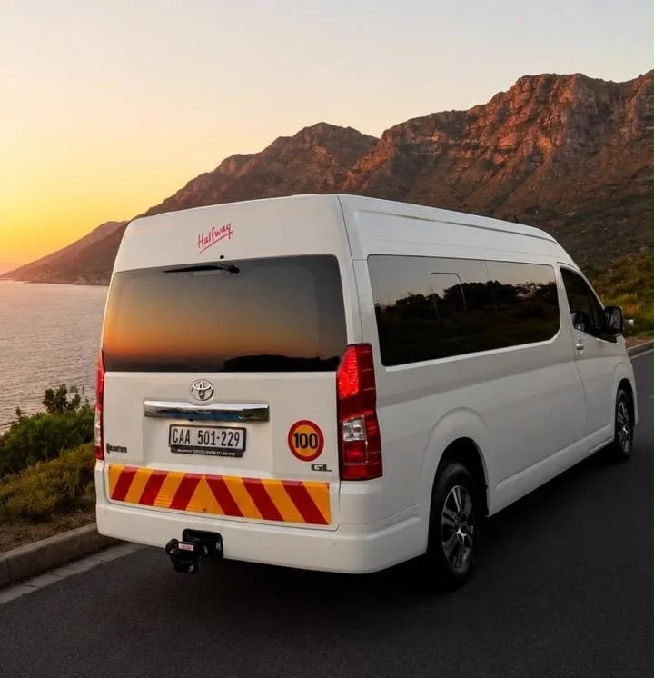 A white Toyota van parked on the side of a scenic road near a body of water with mountains in the background during sunset.