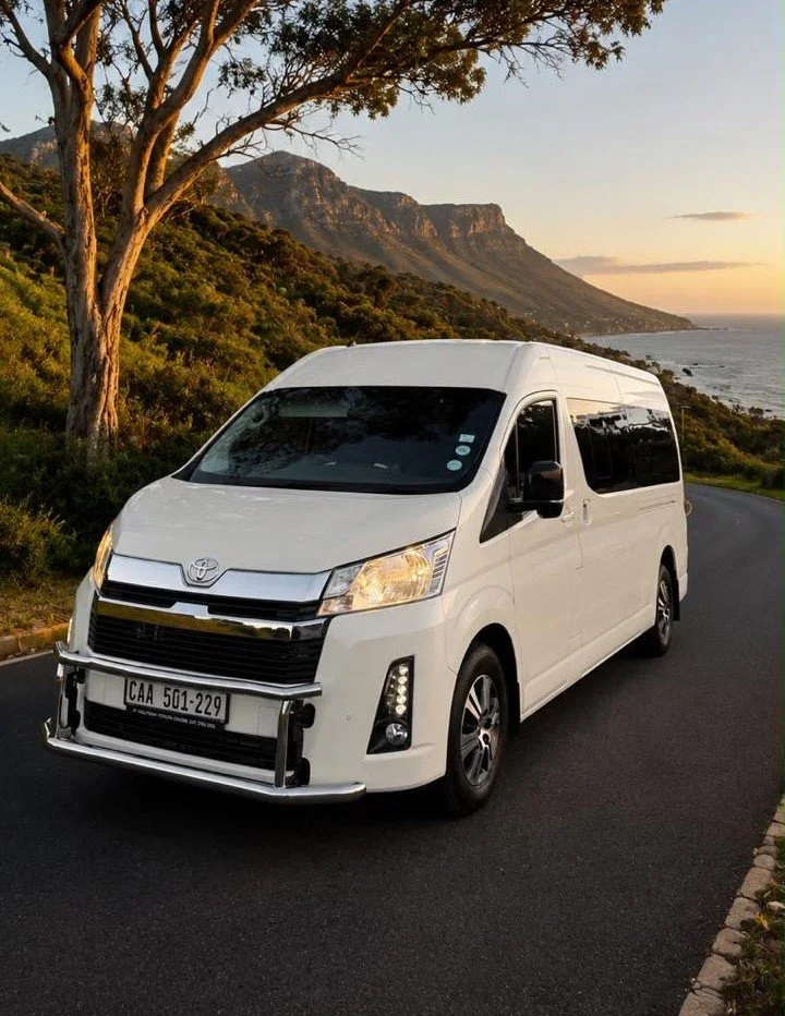 A white Toyota van parked on a scenic coastal road with mountains and ocean in the background during sunset.