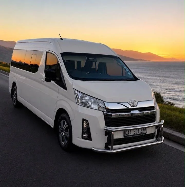 A white Toyota van parked on a road beside the ocean during sunset with mountains in the background.