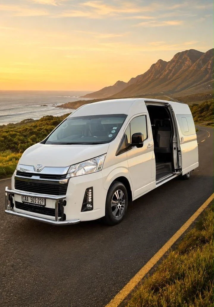 A white cargo van parked on the side of a scenic mountain road during sunset, with the passenger door open revealing the interior.
