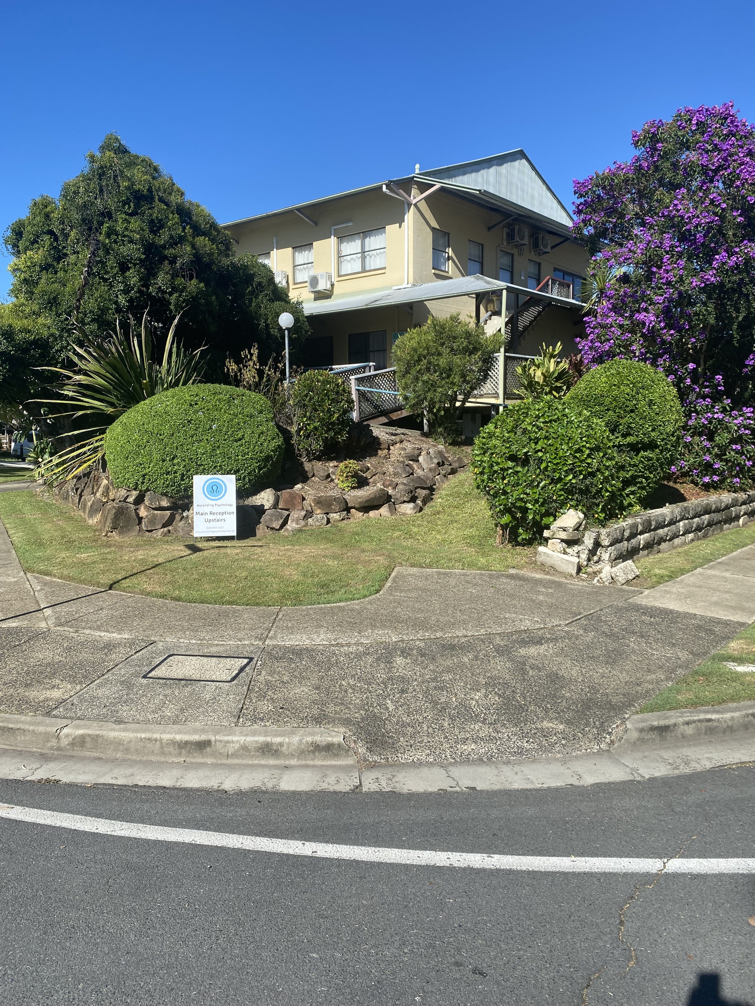 A two-story beige house with multiple windows and air conditioning units, surrounded by lush green plants and a large purple flowering bush; a street curb and sidewalk in the foreground, with a street sign indicating a main reception upstairs.