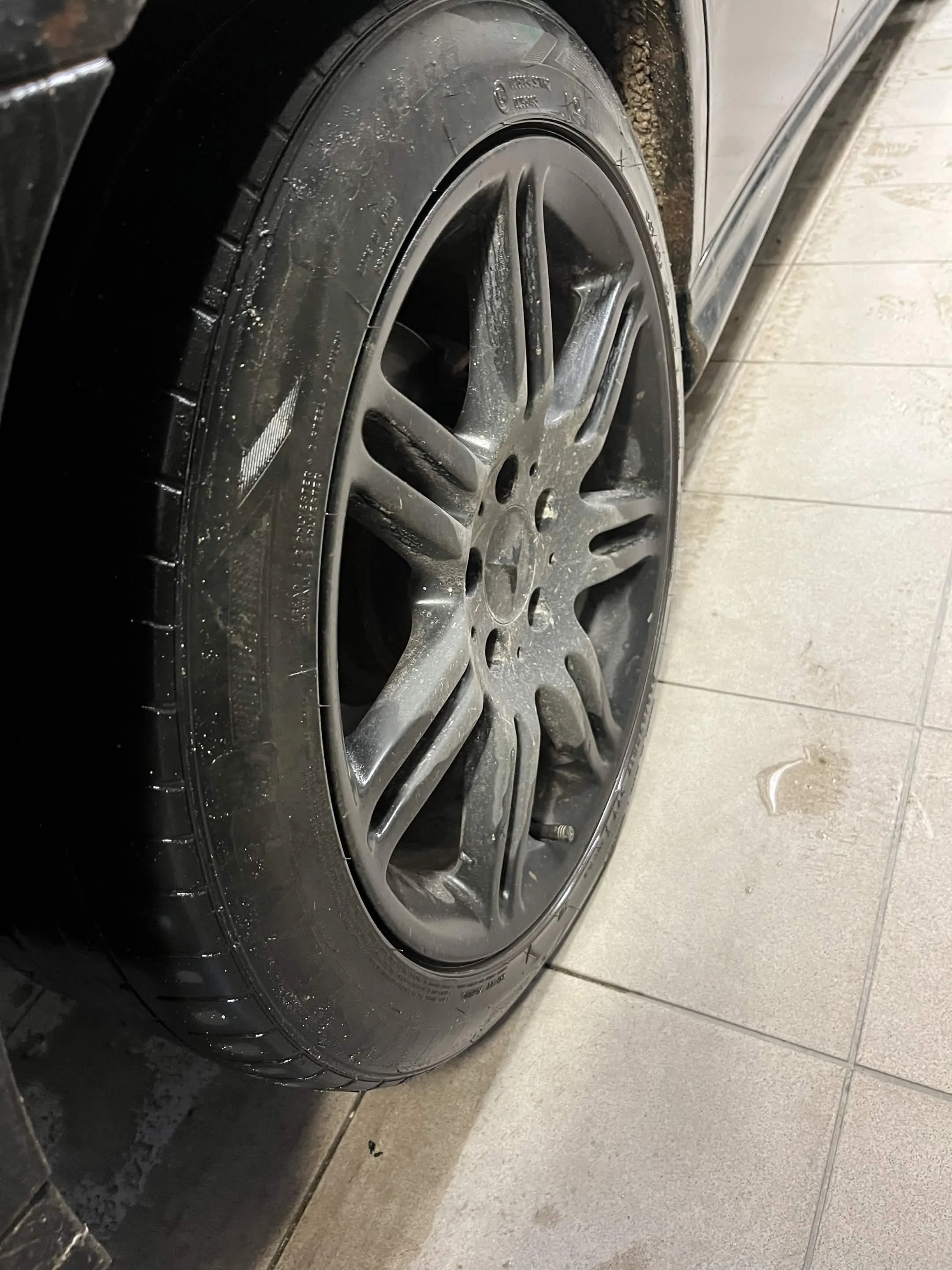 The image shows a close-up of a black car wheel with a silver alloy rim, parked on a tiled floor. The tire appears dusty.