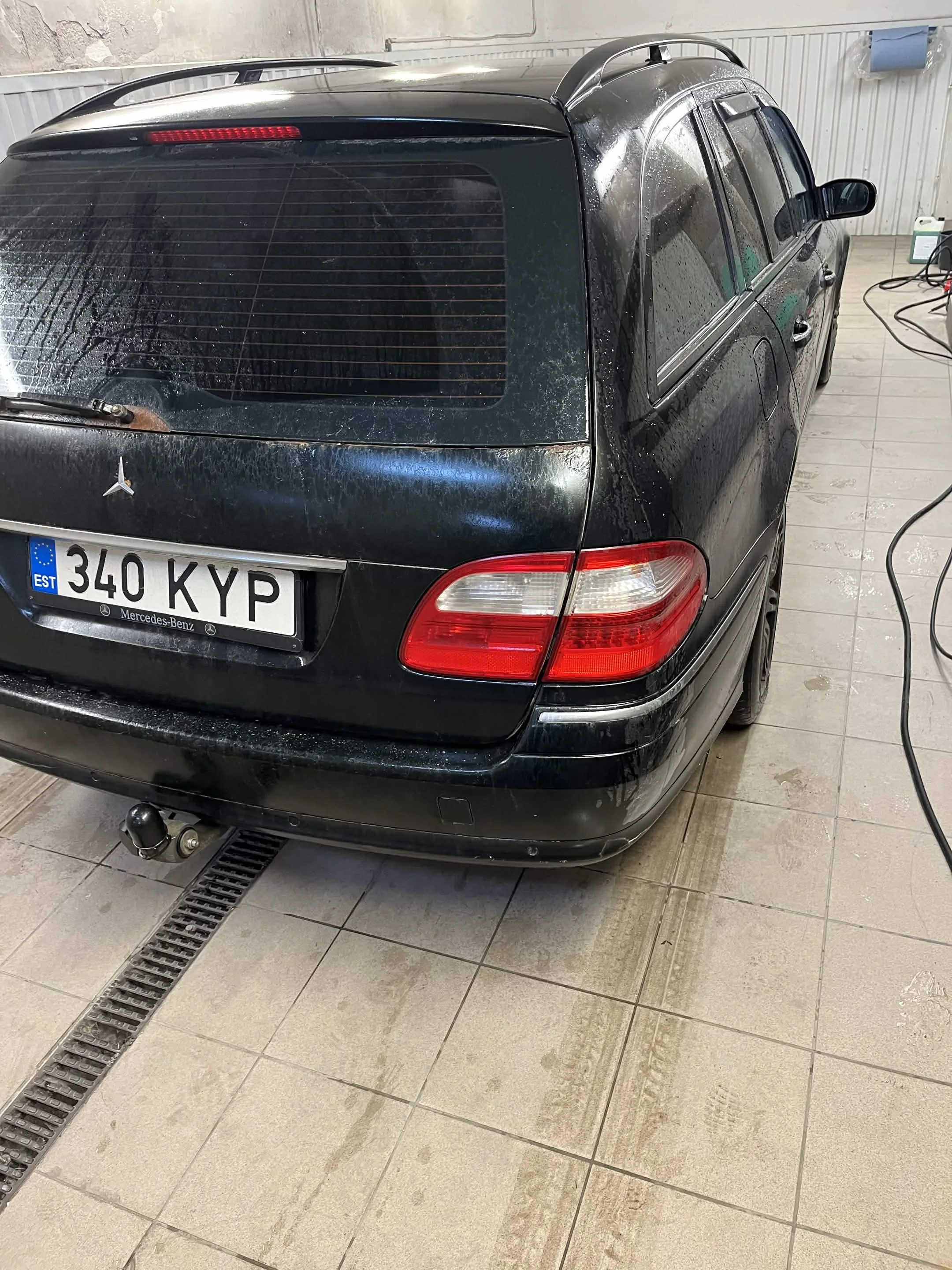 Black Mercedes-Benz station wagon inside a garage or car wash with water droplets on its surface, a European license plate, and cleaning equipment nearby.
