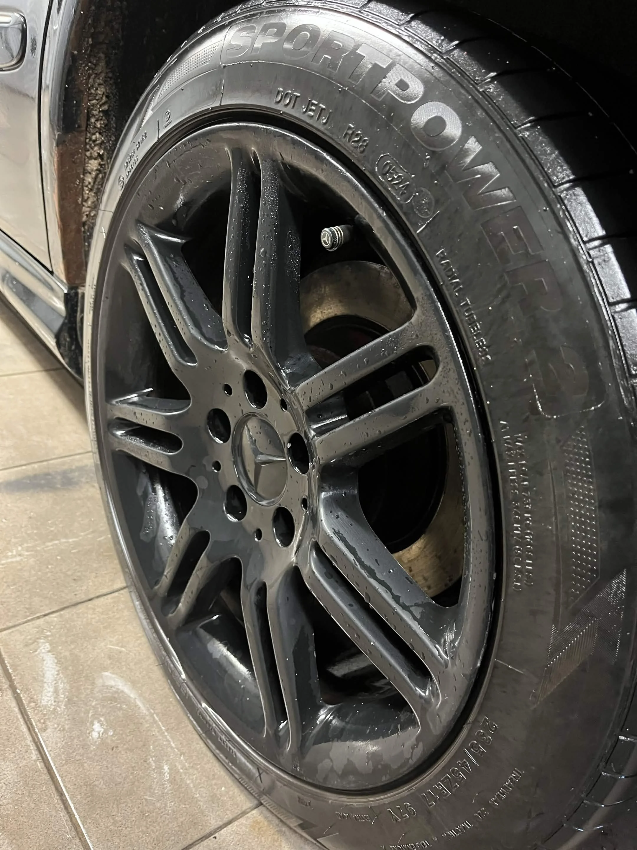 Close-up of a car tire mounted on a shiny alloy wheel, with water droplets and a Mercedes-Benz logo valve cap visible.