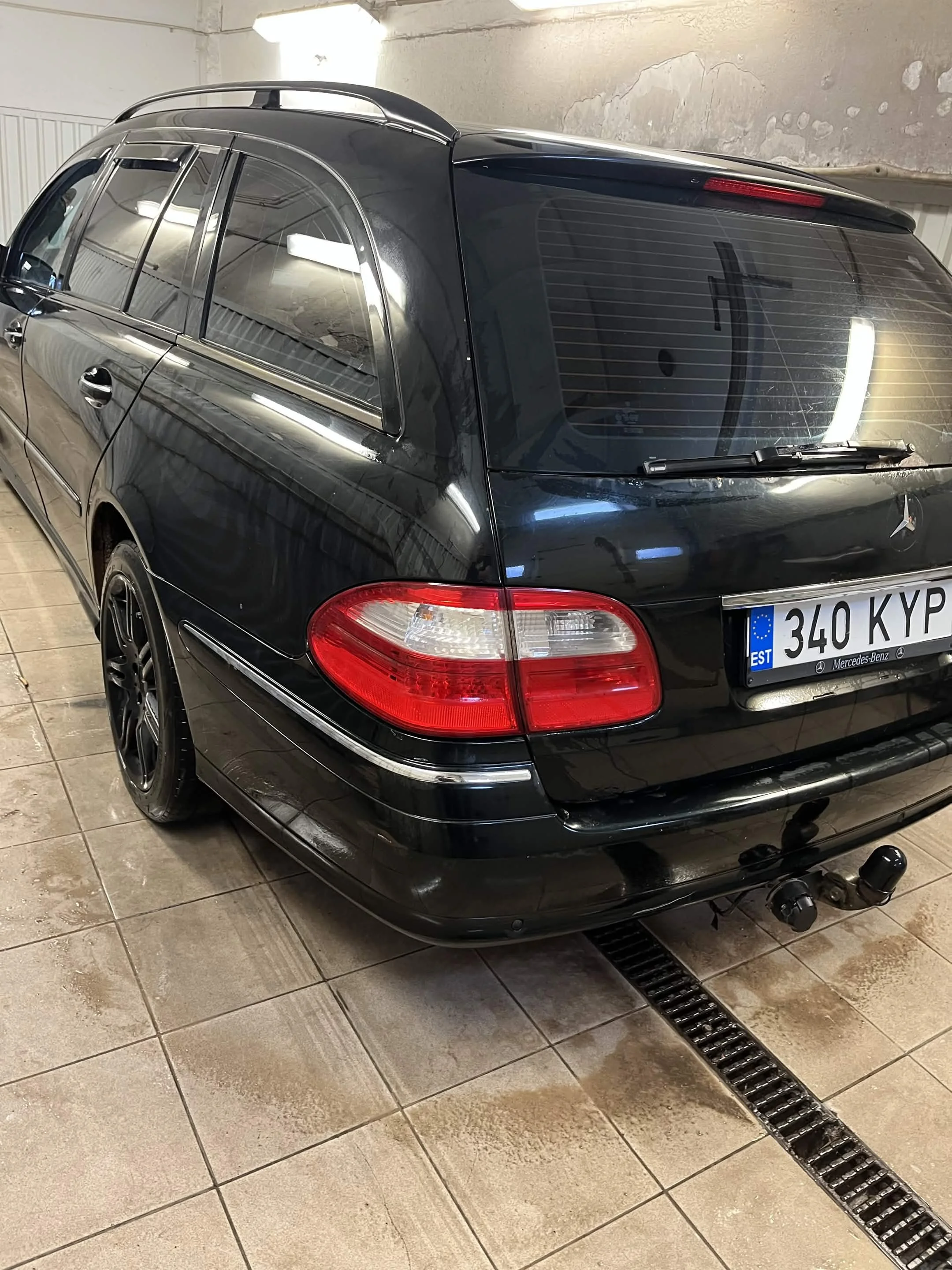 Black Mercedes-Benz station wagon in an indoor garage, photographed from the rear-left side. The car features tinted windows, a roof rack, and a visible European license plate reading '340 KVP,' with a Mercedes-Benz logo and model details.