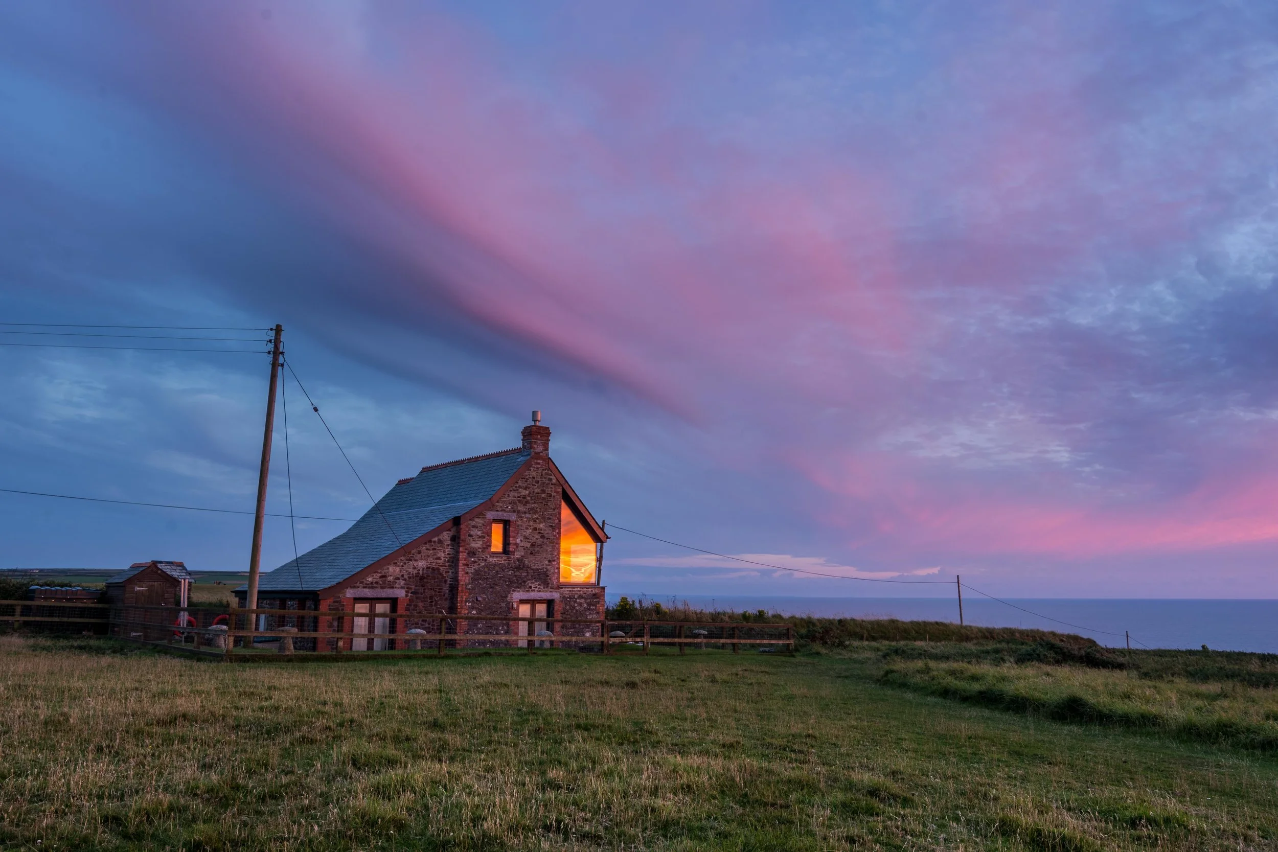 A stone house with a chimney and a lit window, set against a vibrant sky during sunset with purple, pink, and blue clouds. There is a grassy field in the foreground, and power lines and poles near the house.