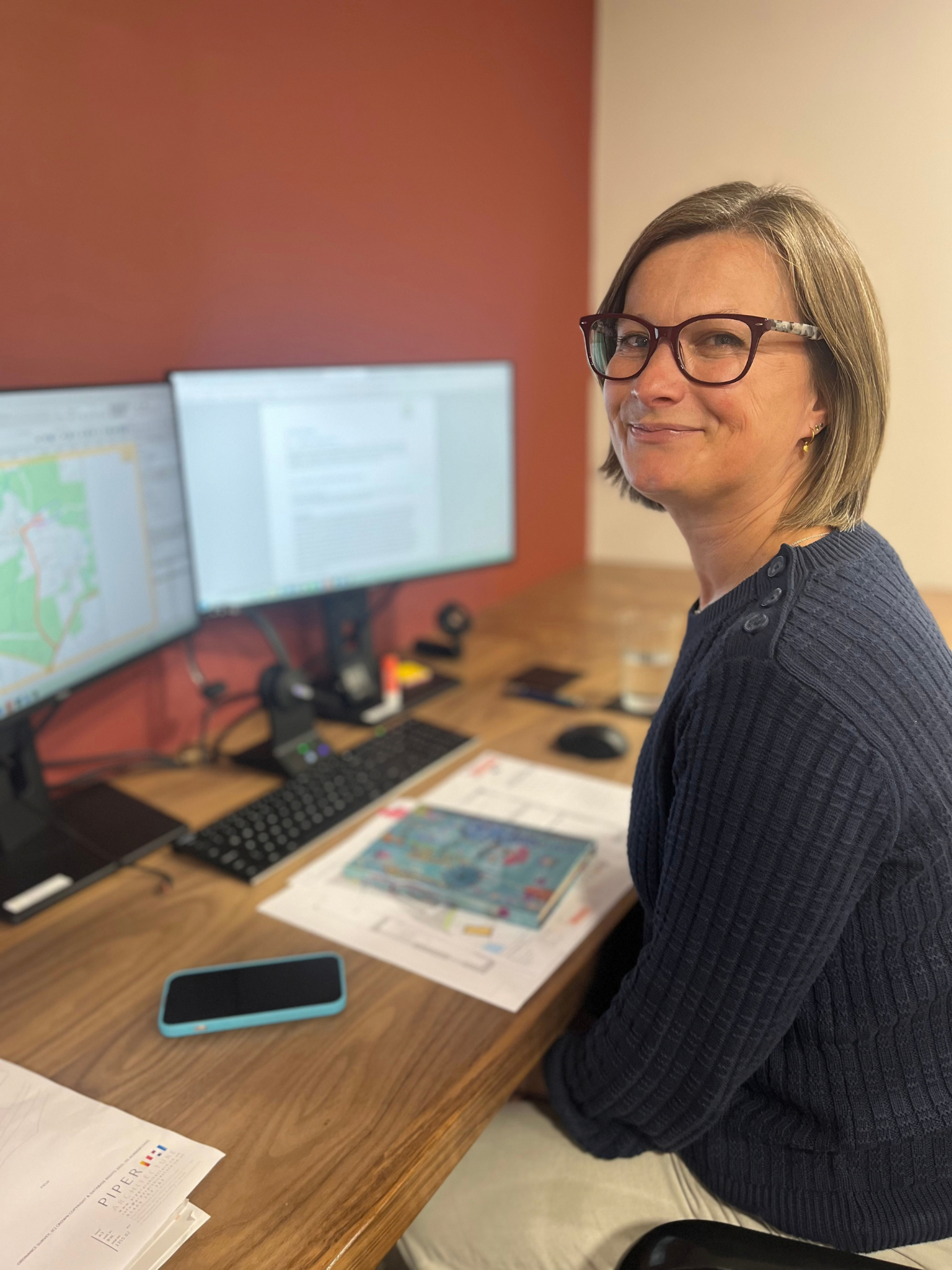 A woman with short brown hair and glasses sitting at a desk with two computer monitors showing a blurred Land Registry map and an architectural fee proposal. On the desk are printed architectural concept design drawings, a notebook and a smartphone.