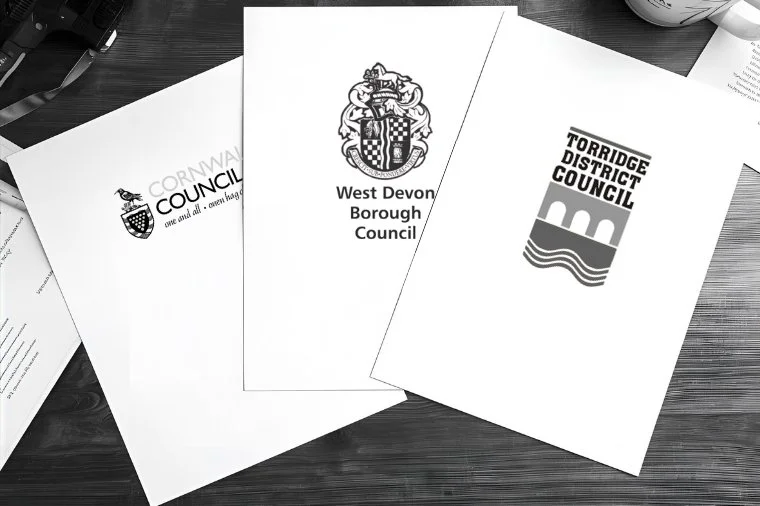 Three official council documents from local government districts in South West England: Cornwall Council, West Devon Borough Council, and Torridge District Council, placed on a wooden surface.