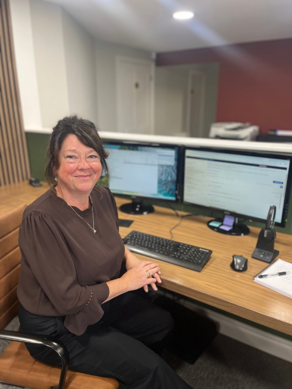 A woman sitting at a desk in an office, smiling, with two computer monitors, a keyboard, a mouse, a notepad, and other office supplies on the desk.