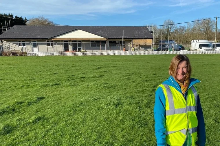 A woman smiling outside on a grassy field, wearing a yellow safety vest and a blue jacket. In the background, there is a building with a dark roof, white fence, and parked vehicles.