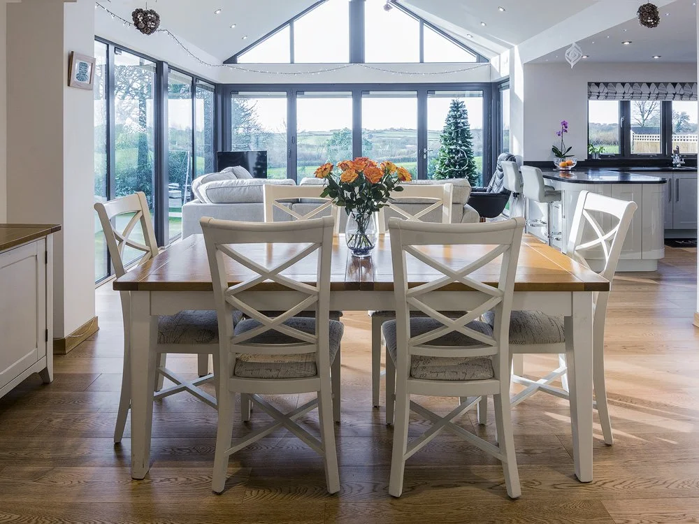 Open-concept dining and living room with large windows, a wooden dining table with six white chairs, a bouquet of orange flowers, a Christmas tree with lights, a white sofa, a flat-screen TV, and a kitchen with white cabinets and bar stools.