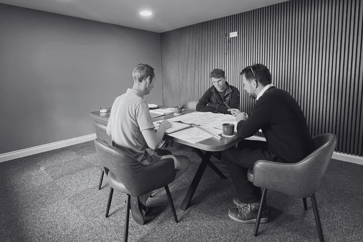 Three men in a meeting room, seated at a table with papers, mugss, and electronic device, engaged in a discussion or work.
