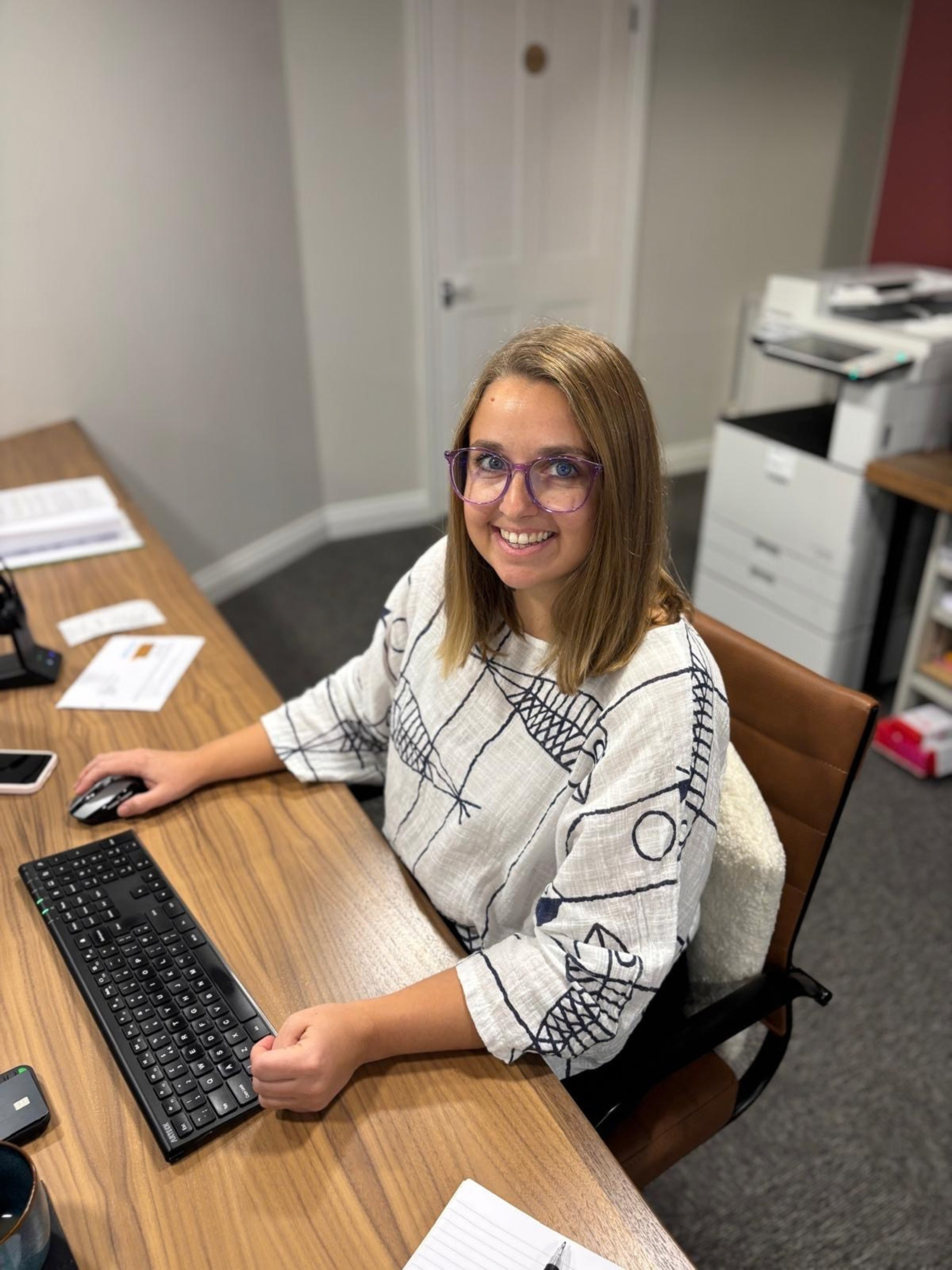 A woman with glasses, long blonde hair, and a white and black patterned blouse sitting at a wooden desk, using a black computer keyboard and mouse, in an office with papers, a notepad, and office equipment around her.