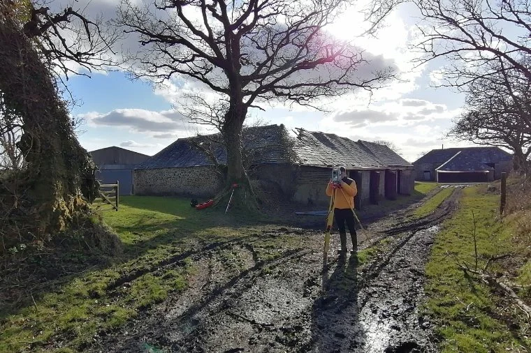 An architectural technician in an orange shirt and dark trousers conducting a topographical survey using a Leica instrument on a tripod outdoors near a rural house, with muddy dirt paths, leafless trees, and a bright sky with clouds.