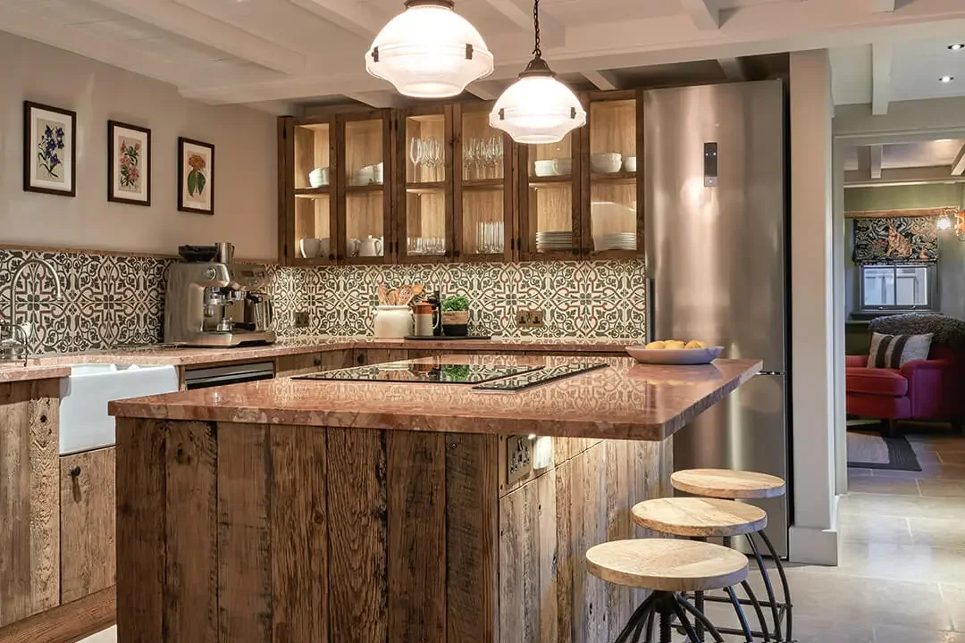 A coy kitchen with wooden cabinets, a pink marble countertop, patterned backsplash tiles, and three white hanging pendant lights. There are three wooden barstools and kitchen appliances, including a coffee machine and a stainless steel refrigerator, with a glimpse of a living room in the background.