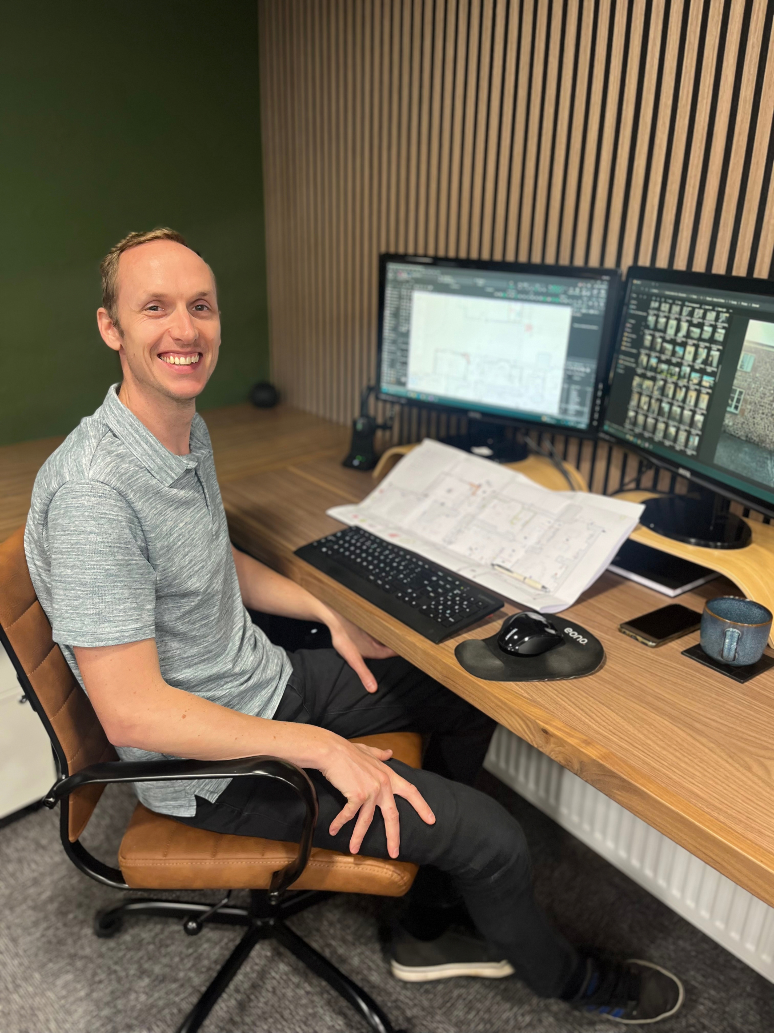 A smiling man with short hair in a grey t-shirt sitting at a wooden desk with two computer screens showing detailed technical architectural plans and site survey photographs. On the desk is a keyboard, mouse, mug, smartphone, and a printed drawing.