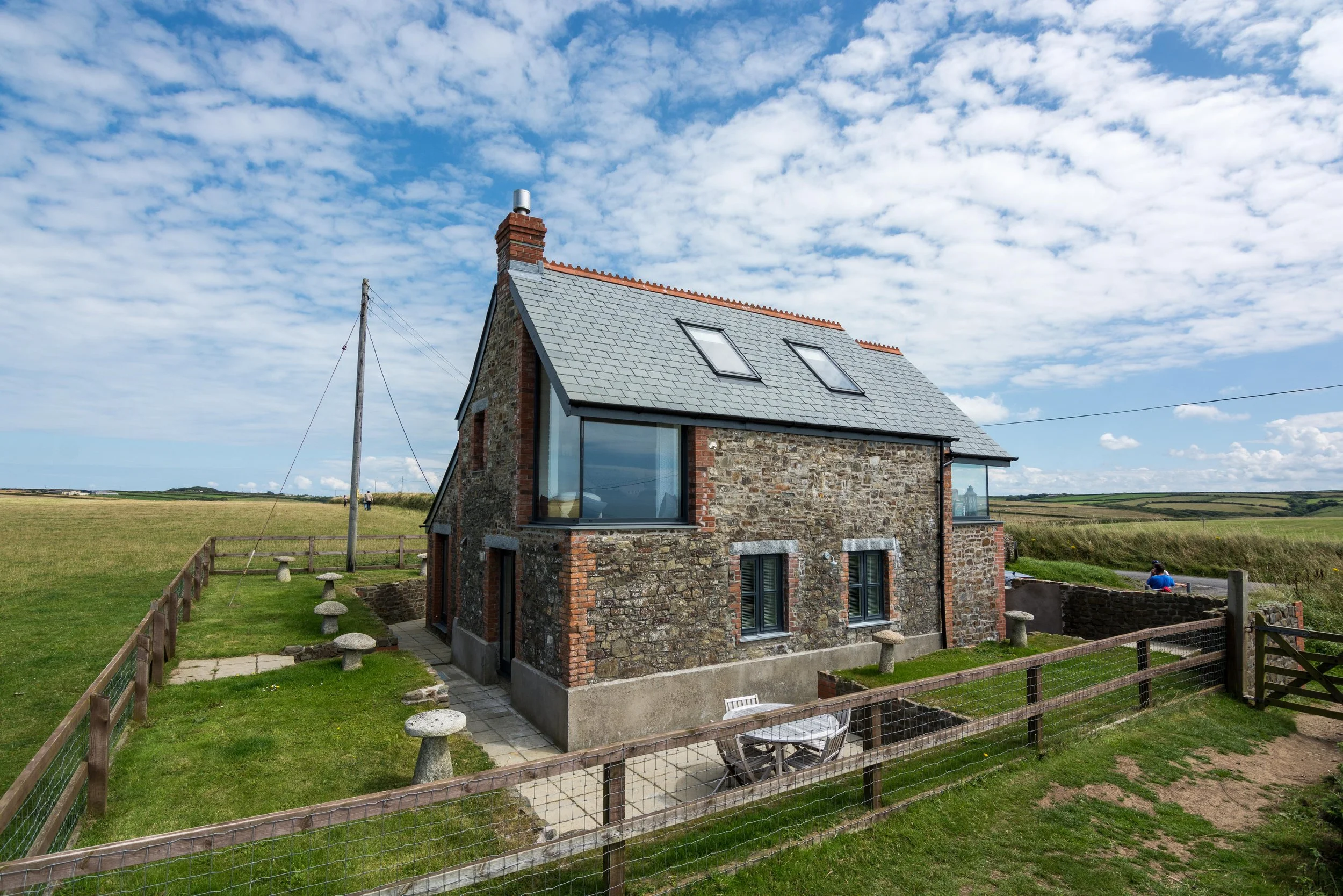 A stone house with a gray tiled roof and skylights, surrounded by a fenced yard with outdoor furniture, set in a rural landscape under a partly cloudy sky.