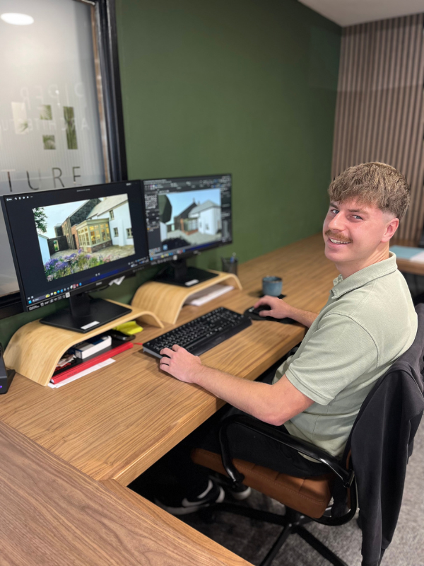 A young man with an impressive moustache sitting at a wooden desk using a computer with dual monitors, editing 3D concept design renders, with a green wall in the background.