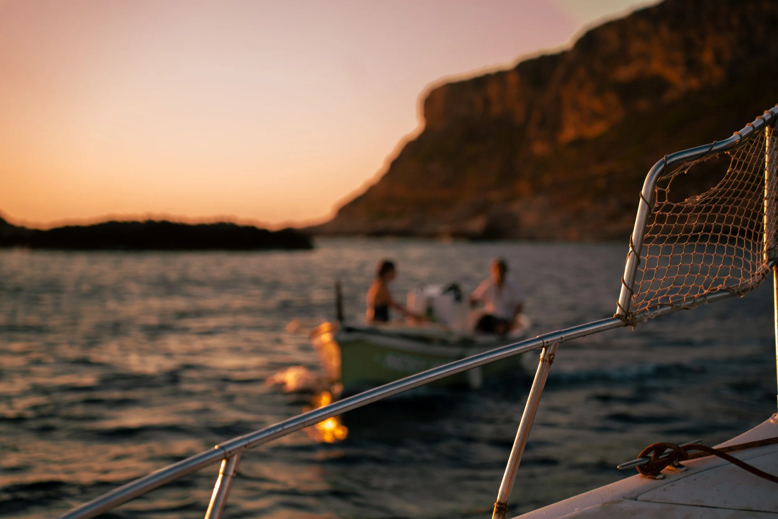 Vista di un barbecue con il reticolato e il tubo di metallo in primo piano, sullo sfondo un mare al tramonto con due persone che parlano su una barca, e una montagna sulla destra.