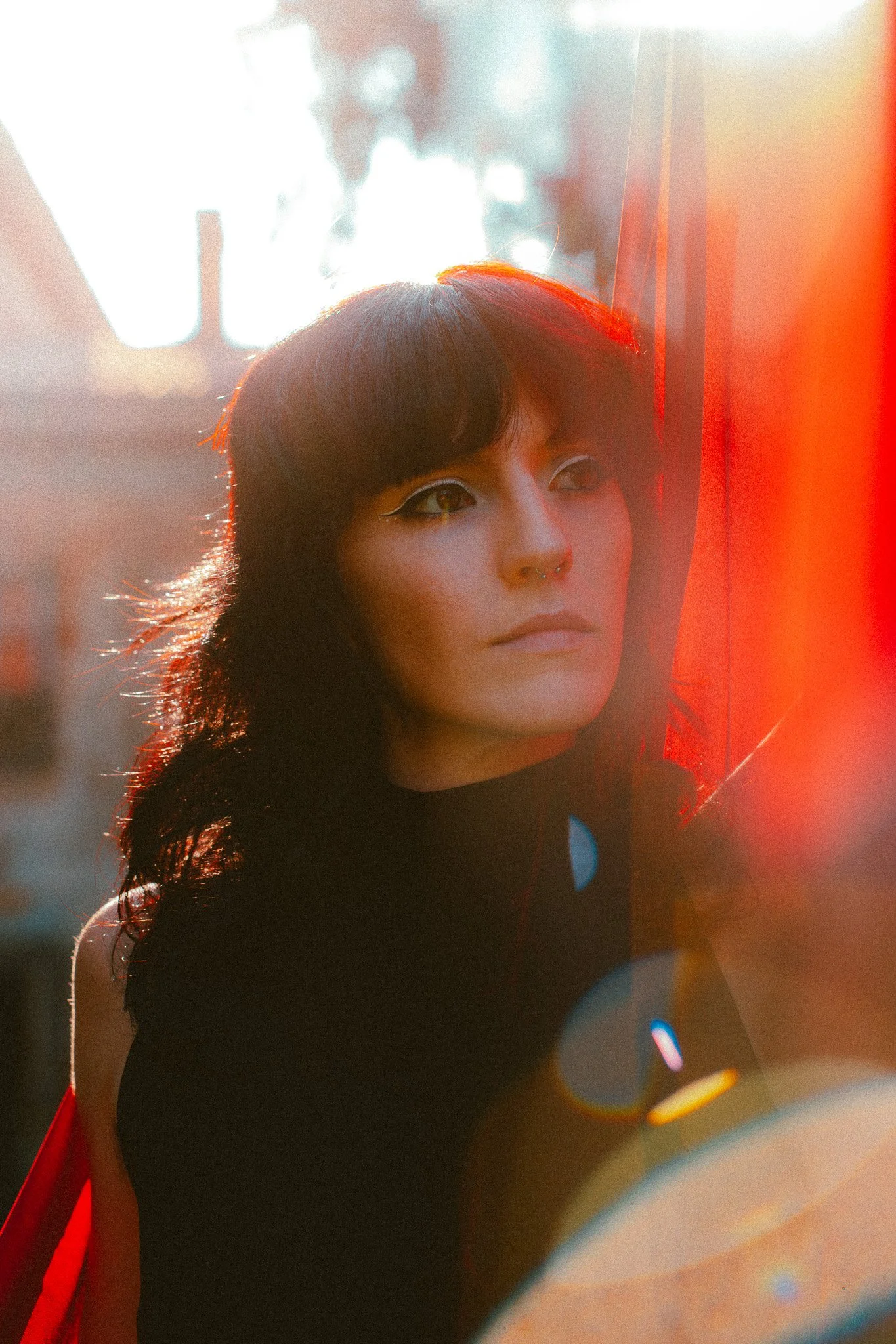 Ritratto di una donna con trucco marcato, cappello nero e capelli ricci, illuminata dal sole con effetti di luce colorata sullo sfondo.