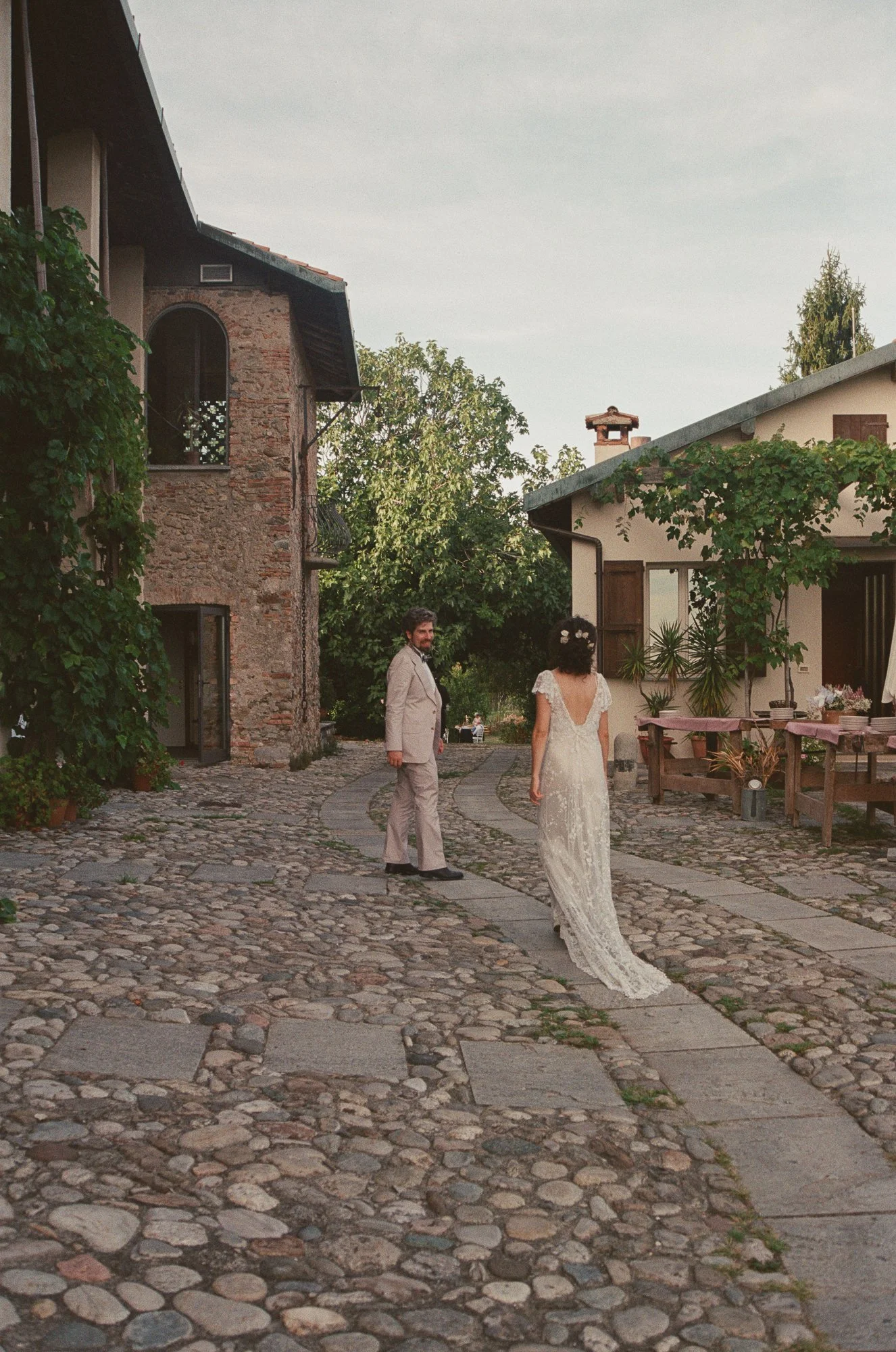 Una coppia di sposi si tiene per mano all'aperto in un cortile in pietra. La donna indossa un abito da sposa lungo con un cappello di fiori, l'uomo un completo beige. Sullo sfondo ci sono case di campagna e alberi verdi.