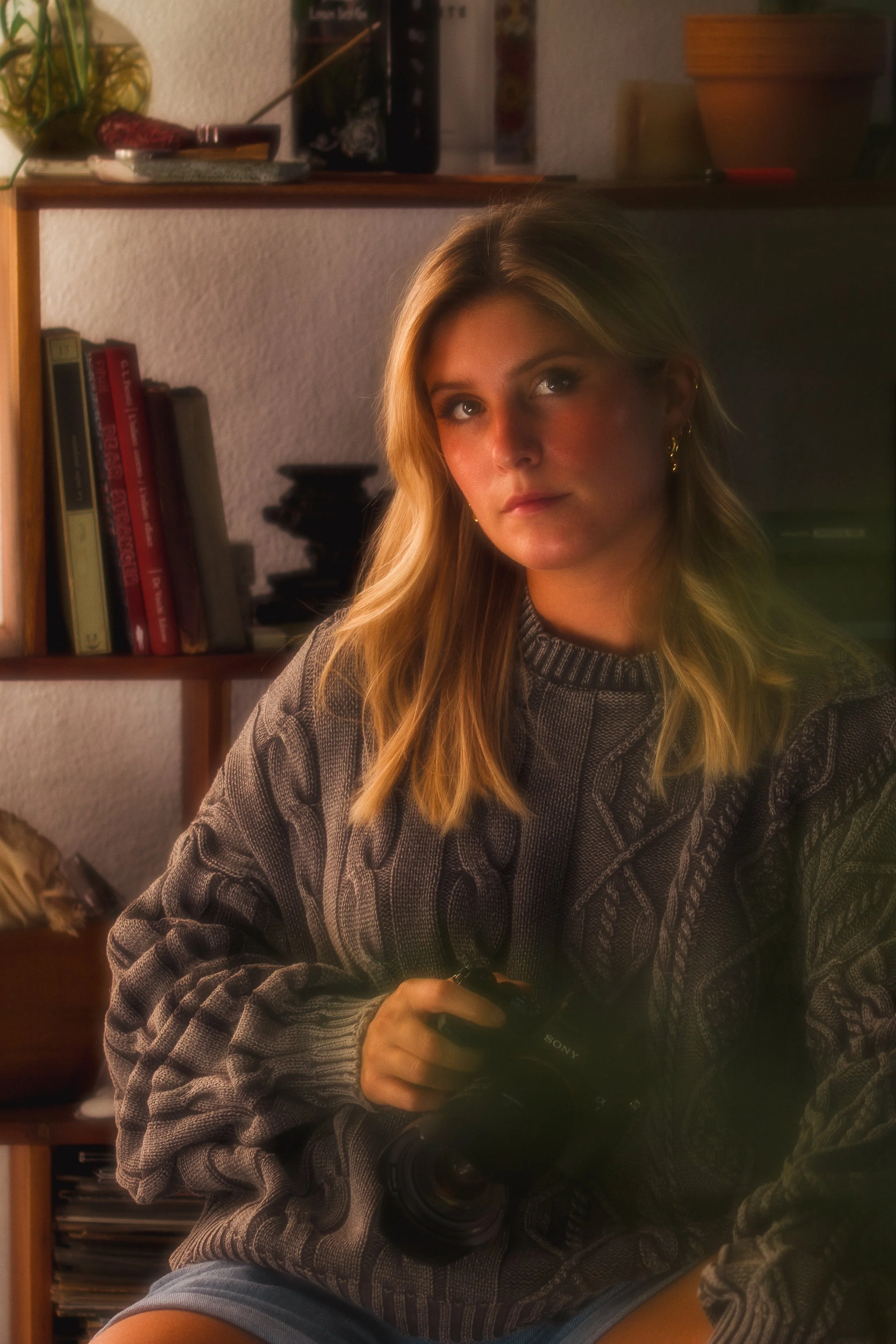 Ragazza con lunghi capelli biondi seduta in una stanza con scaffali pieni di libri, che tiene in mano una fotocamera Sony