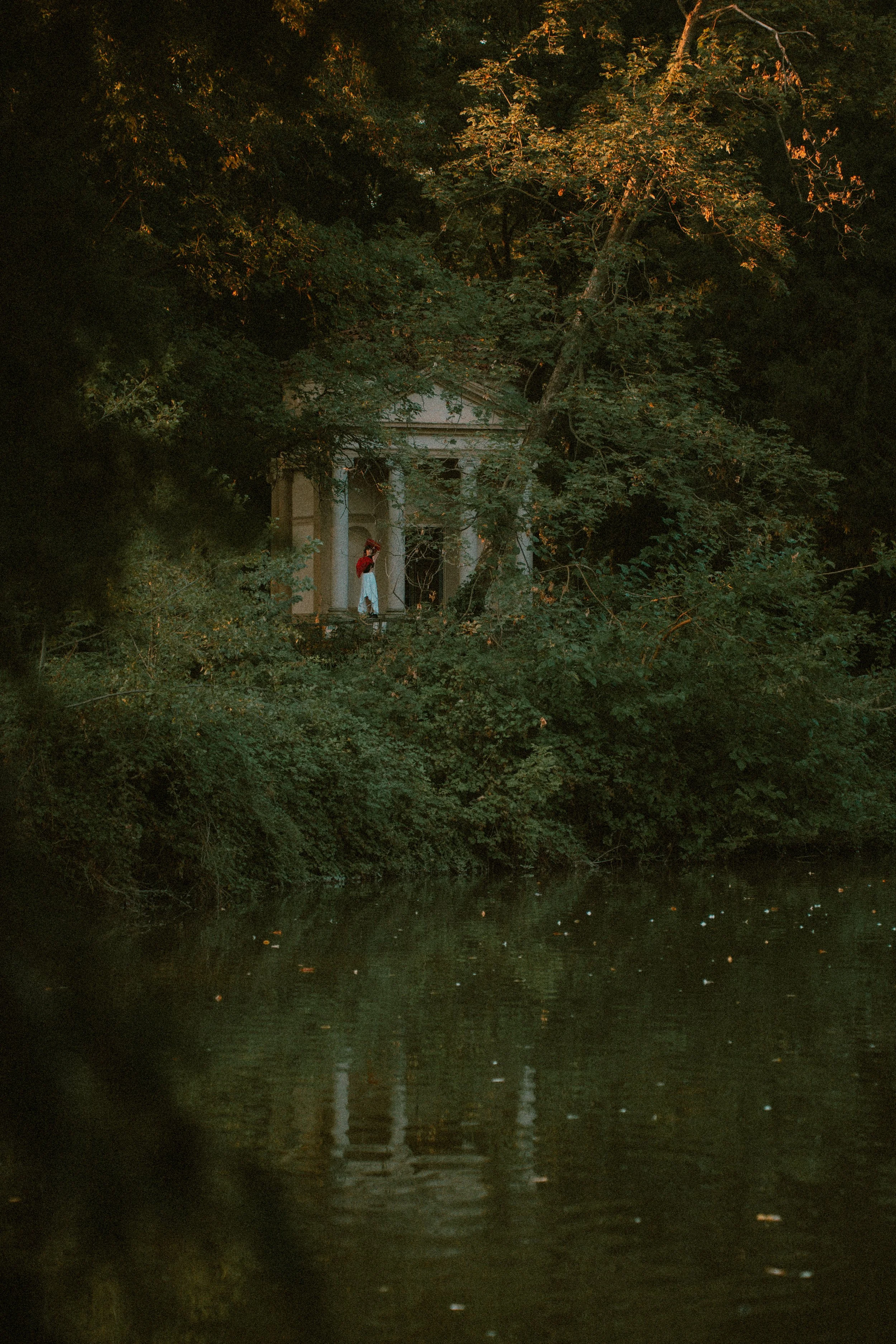 Una donna con abbigliamento vintage, cappello rosso e gonna lunga, si trova di fronte a un edificio antico circondato da un bosco, vicino a un fiume o lago, durante il tramonto o l'alba.