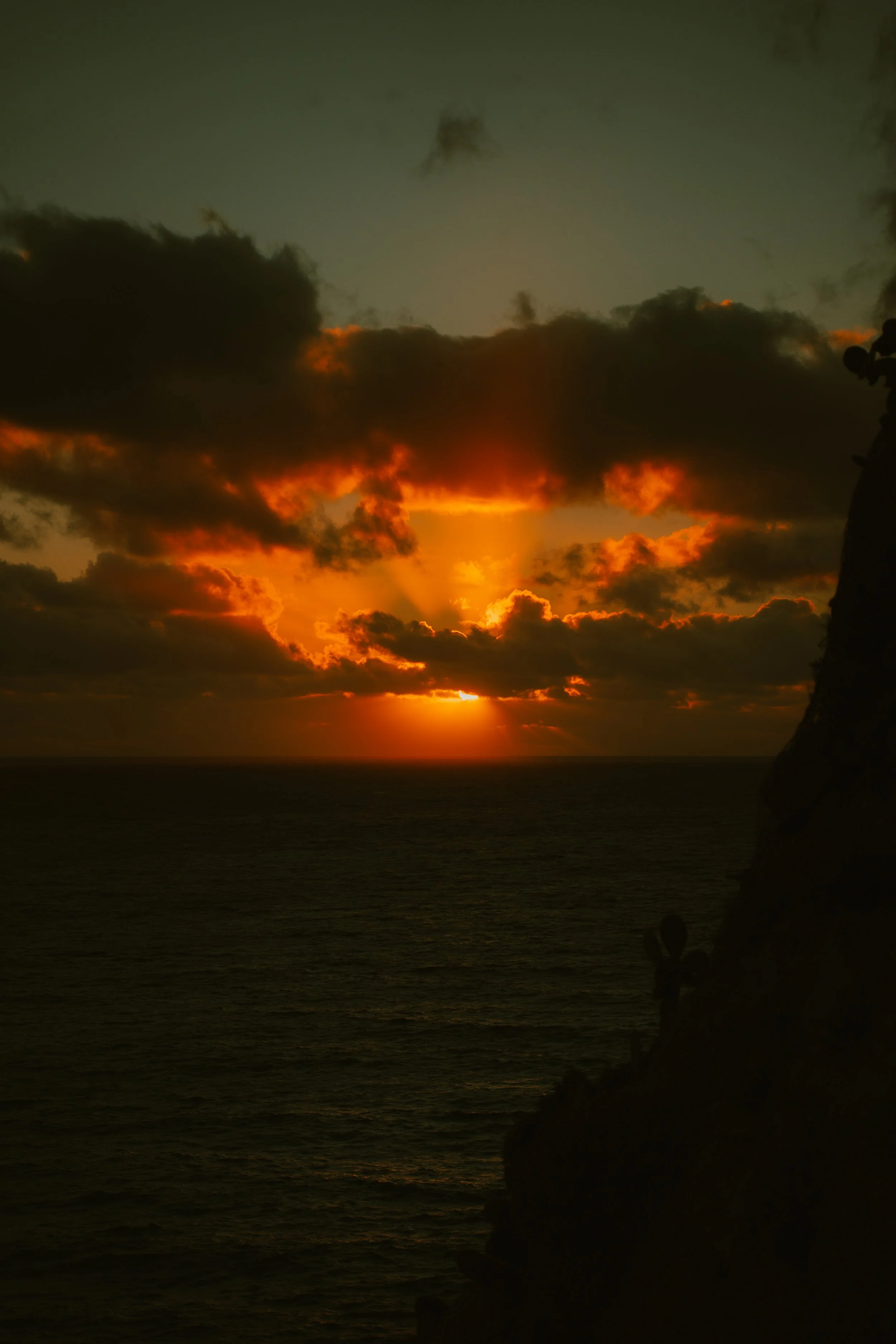 Tramonto sul mare con nuvole scure e raggi di sole che filtrano dietro di esse, vista dalla costa con una scogliera a destra e alcune persone in lontananza.