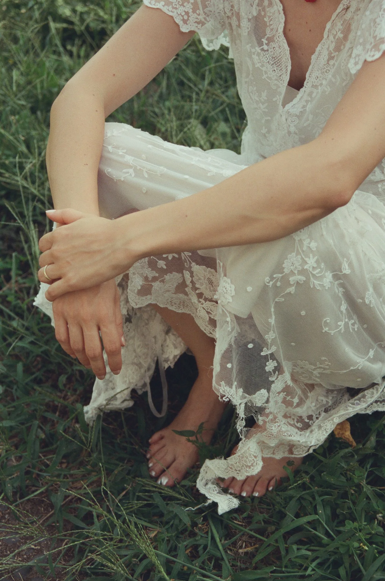 Una donna indossa un vestito bianco in pizzo con dettagli floreali, seduta sull'erba con i piedi nudi, le sue mani sono raccolte davanti a lei.