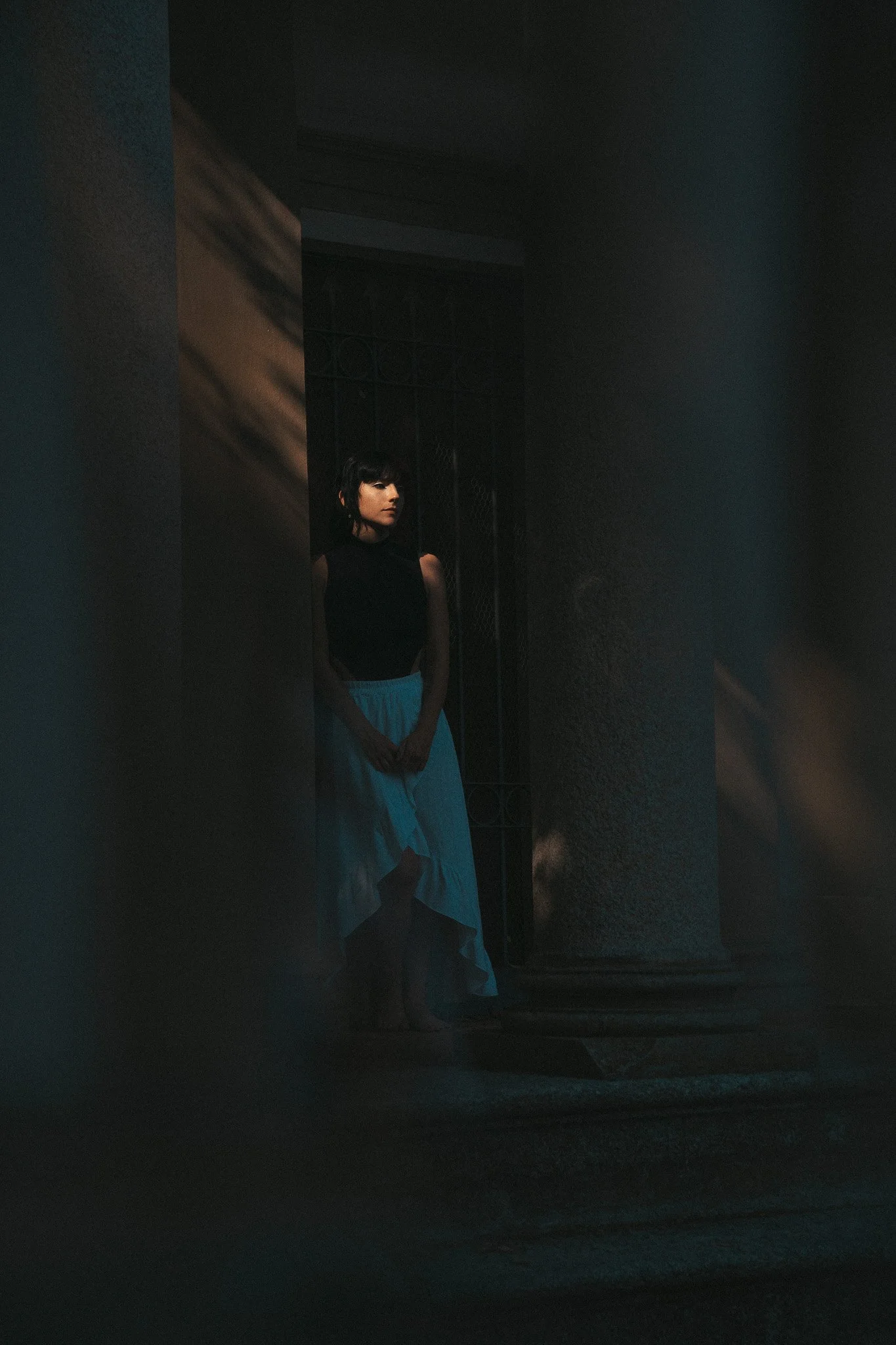 Una donna vestita con una gonna lunga e un top nero si trova in un ambiente scuro, con luce che illumina parzialmente il suo volto e il suo corpo, creando un'atmosfera misteriosa.