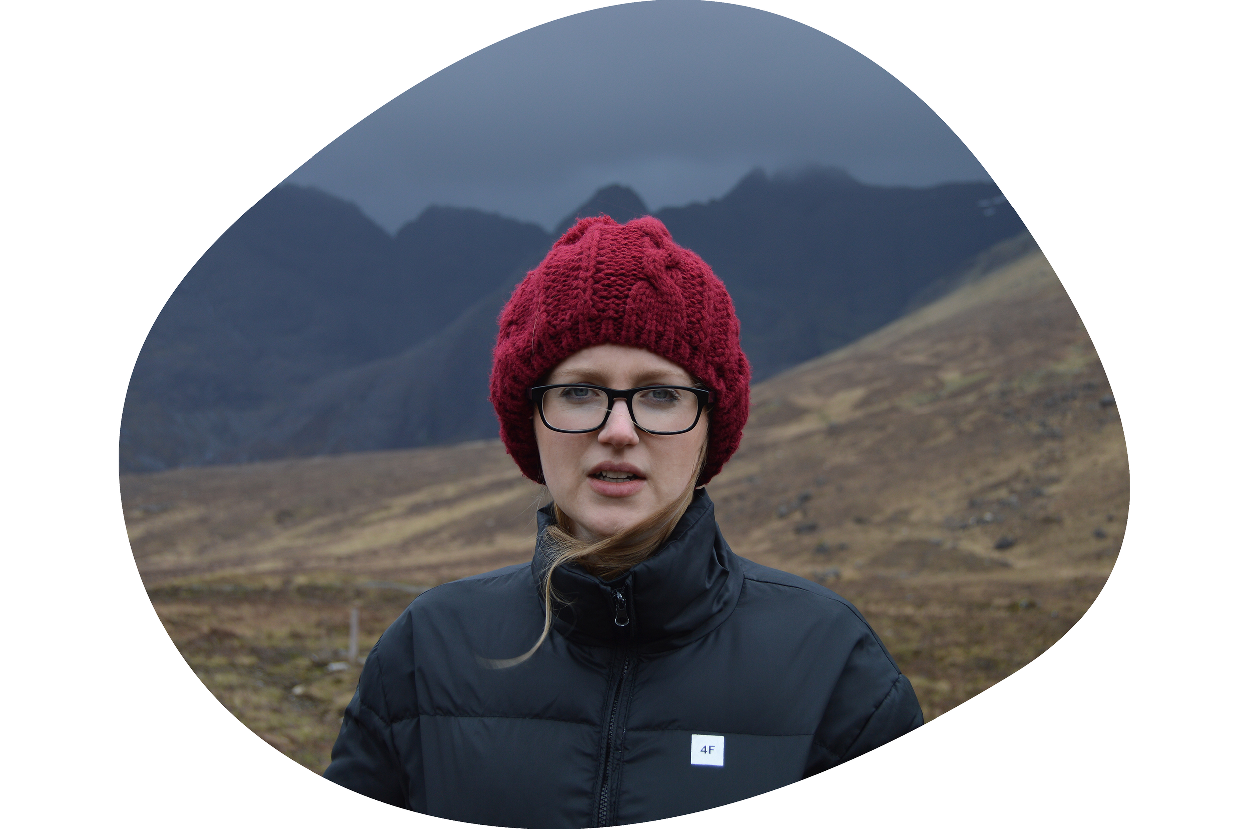 A woman wearing glasses, a red knit beanie, and a black jacket standing outdoors in a mountainous landscape with gray clouds in the sky.