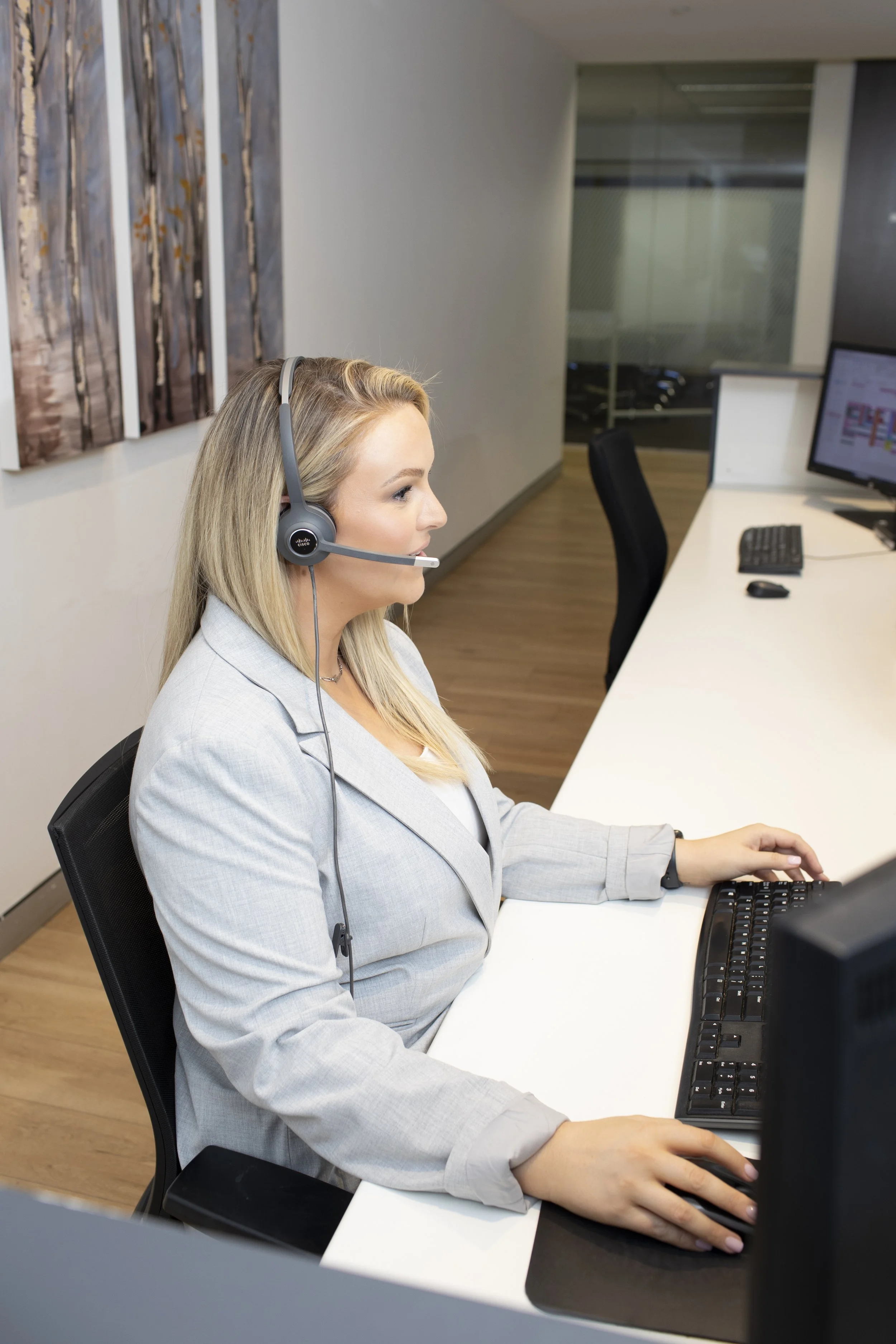 A woman with blonde hair wearing a gray blazer and headset sitting at a white desk in an office, working on a computer.