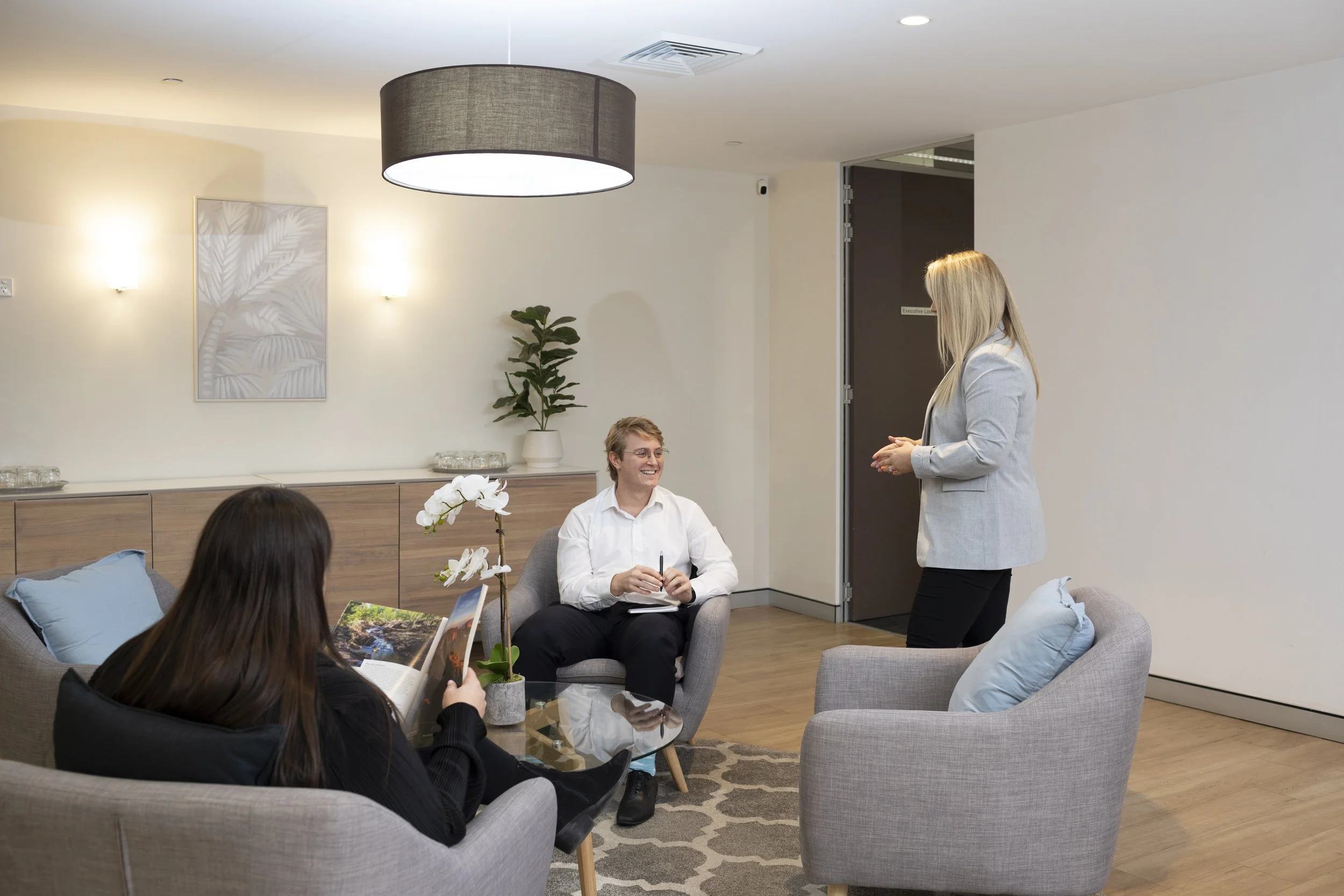 A group of four people conversing in a modern office reception at 115 Pitt St. One woman stands talking to two seated individuals. The room features light-colored walls, a wooden cupboard, decorative art, a plant, and comfortable chairs with pillows.