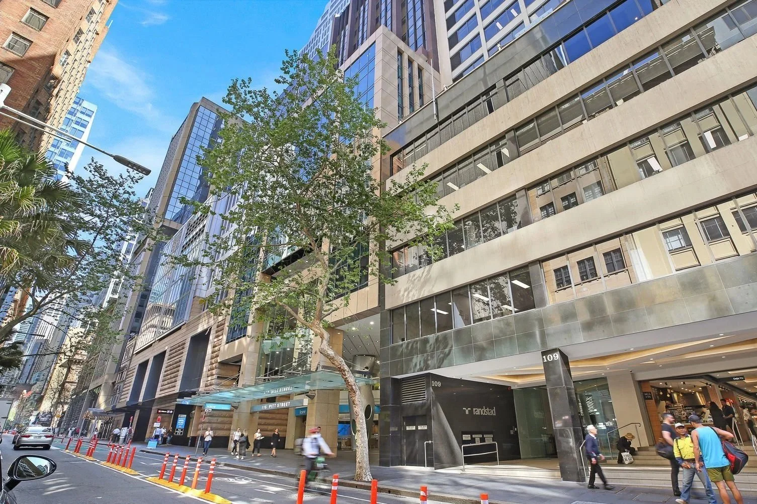 Street view of a modern urban area with tall glass and concrete buildings, trees, and pedestrians walking on the sidewalk. There are orange traffic cones and a bike lane in the foreground, and a hotel entrance with the address 109 and a Randstad office visible.