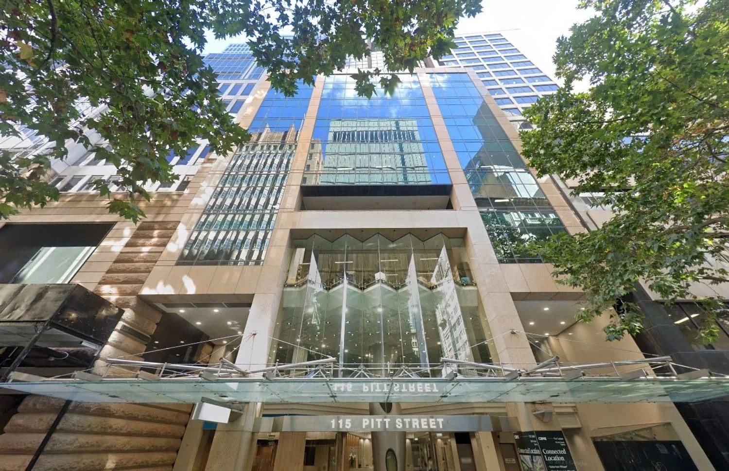 View of a tall modern office building with glass windows reflecting the sky and surrounding buildings, located at 115 Pitt Street, framed by green trees at the street level.