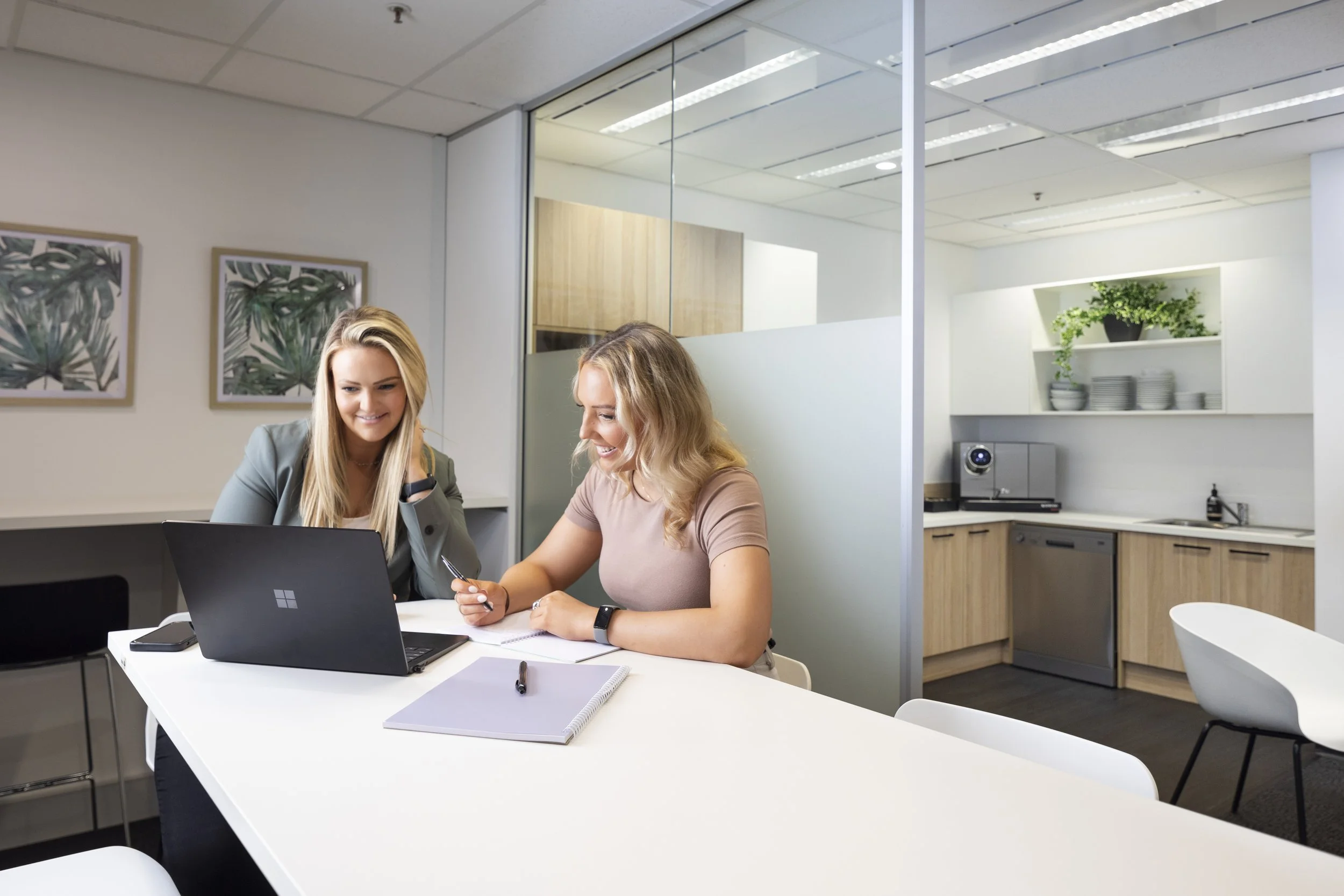 Two women are sitting at a white table in a modern office kitchen area, smiling and engaging with a laptop. The room has white walls, a coffee machine, functional cabinets, and decorative plant art on the wall.